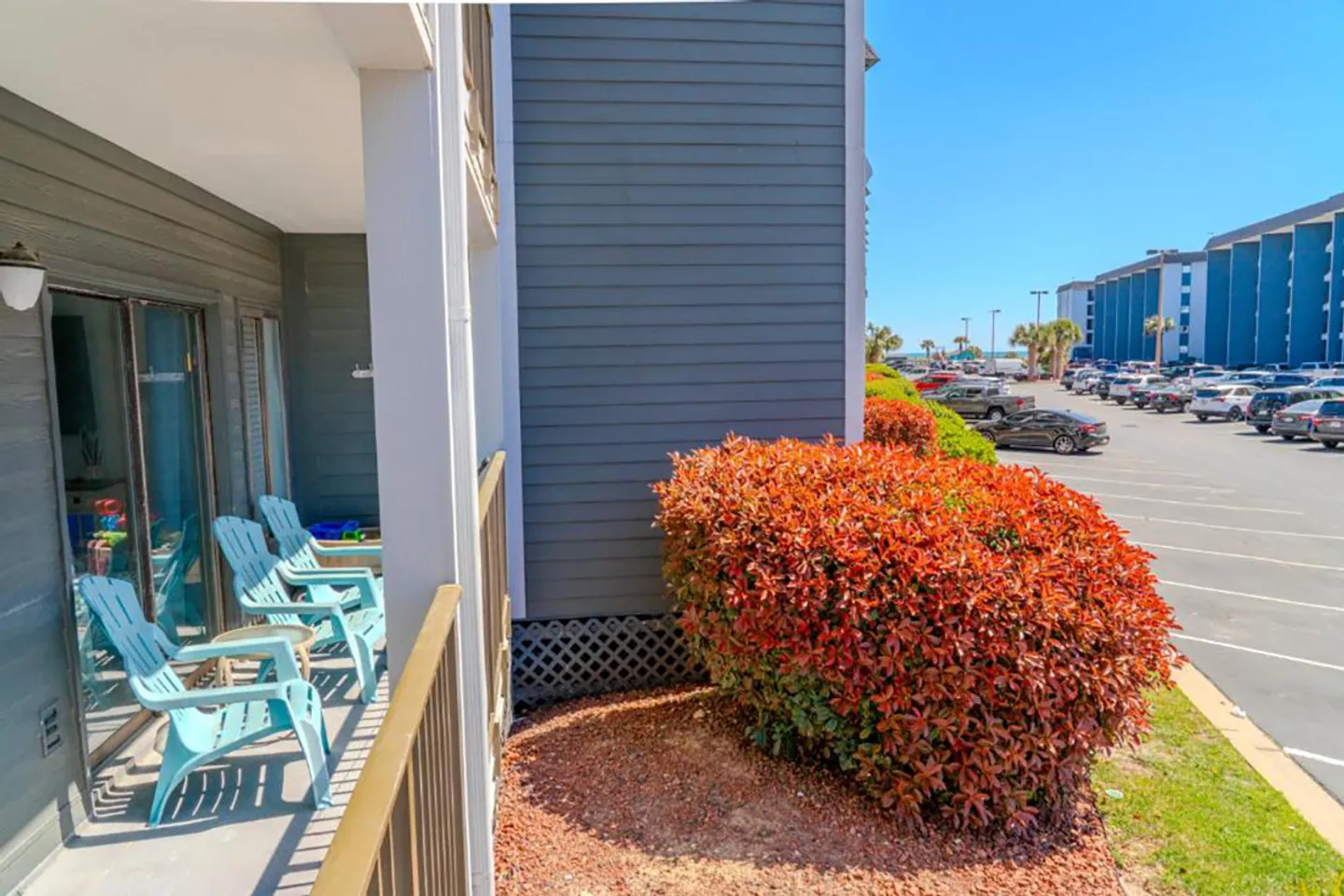 Ground floor covered balcony with light blue chairs next to landscaping and a parking lot.