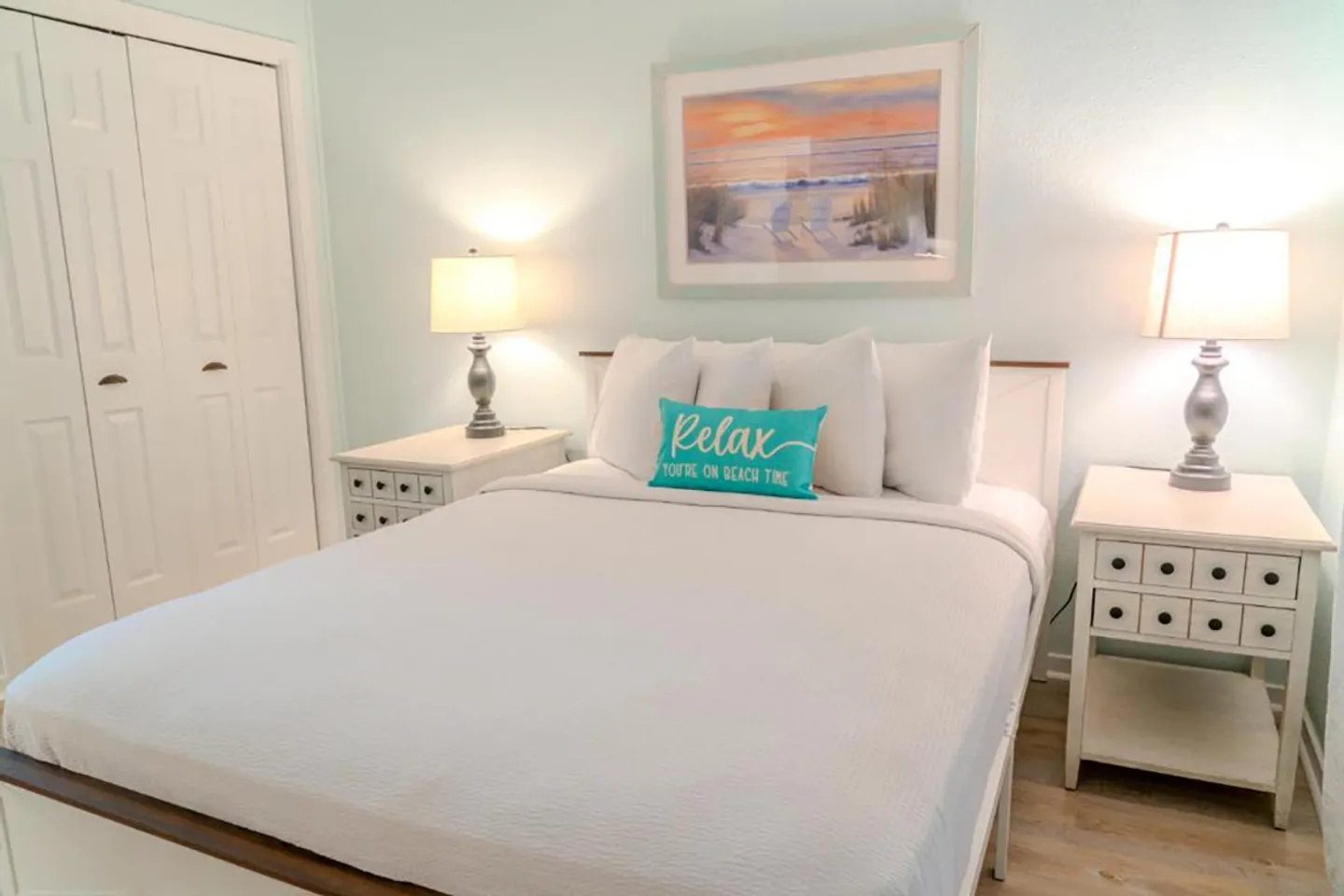 Bedroom with a white-dressed bed flanked by matching nightstands with lamps and framed coastal art on the pale blue wall.