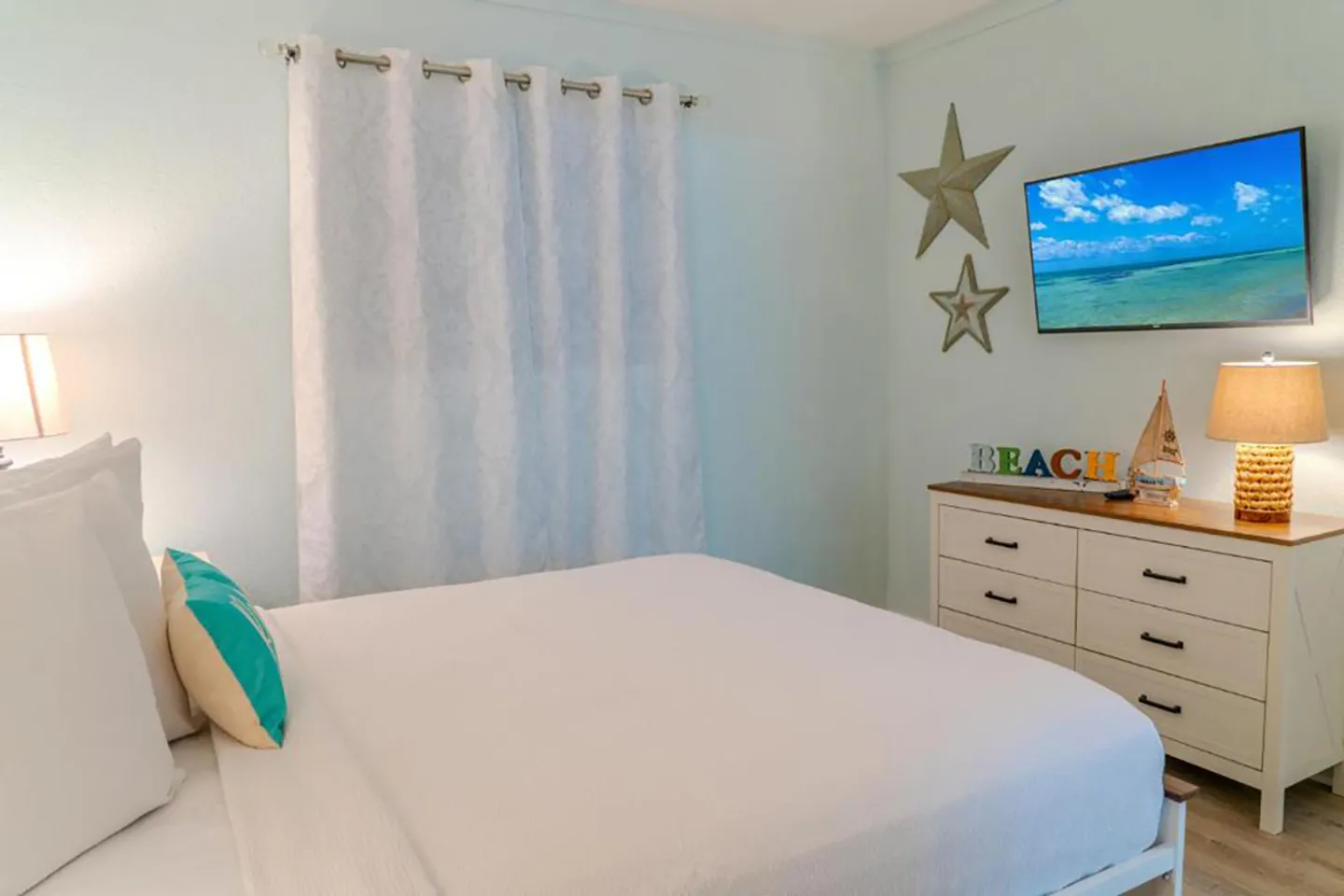 Bedroom with a white-dressed bed, window curtain, dresser, and wall-mounted television.