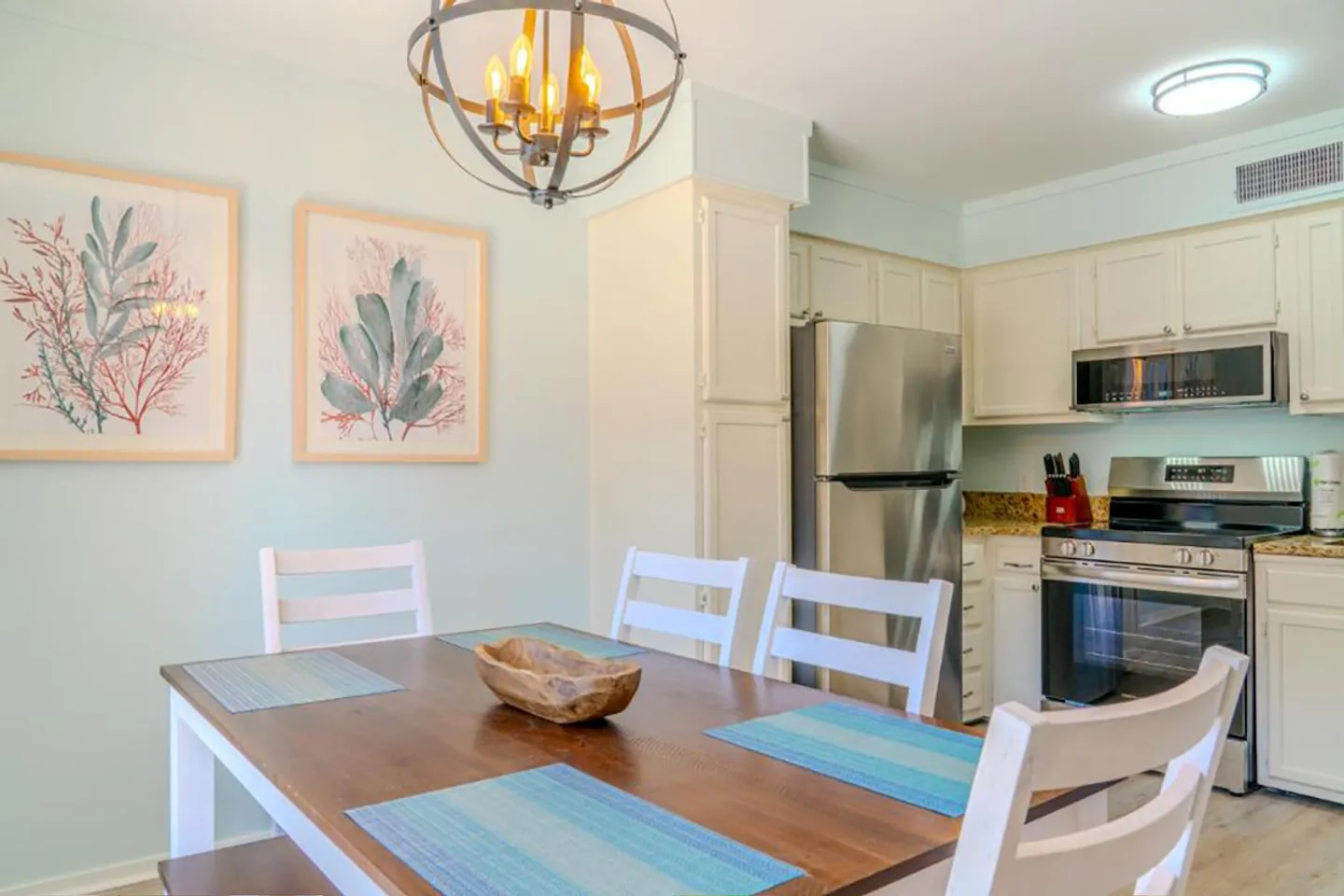 Dining area with a wooden table, white chairs, and an orb chandelier, adjacent to a kitchen with stainless steel appliances.