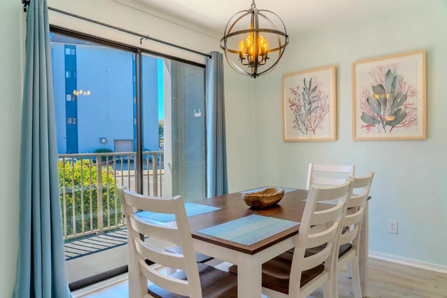 Dining area with a wooden table, white chairs, orb chandelier, and sliding glass doors leading to a balcony.