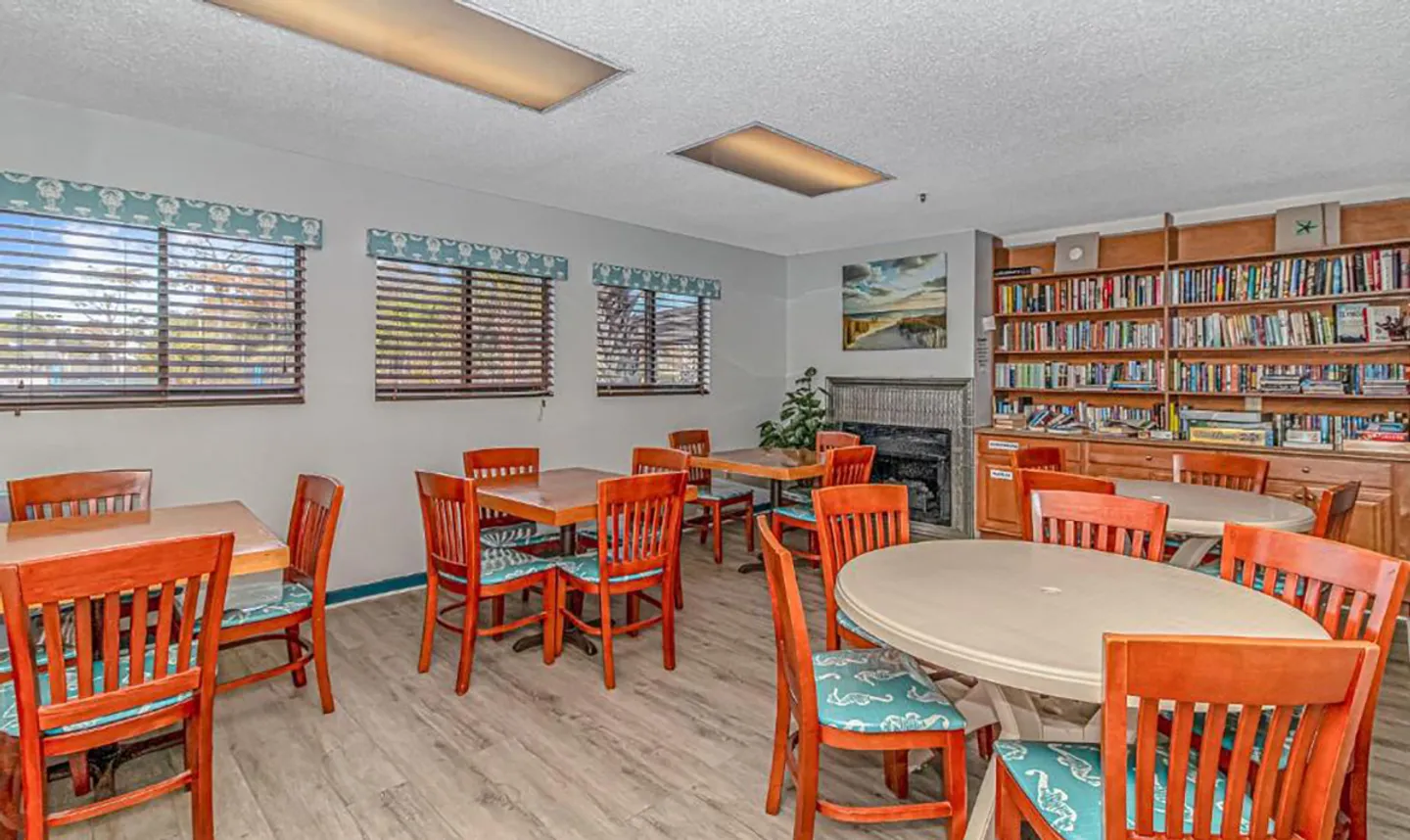 Community room with multiple dining tables, wooden chairs with blue patterned seats, windows with blinds, a fireplace, and built-in bookshelves filled with books.
