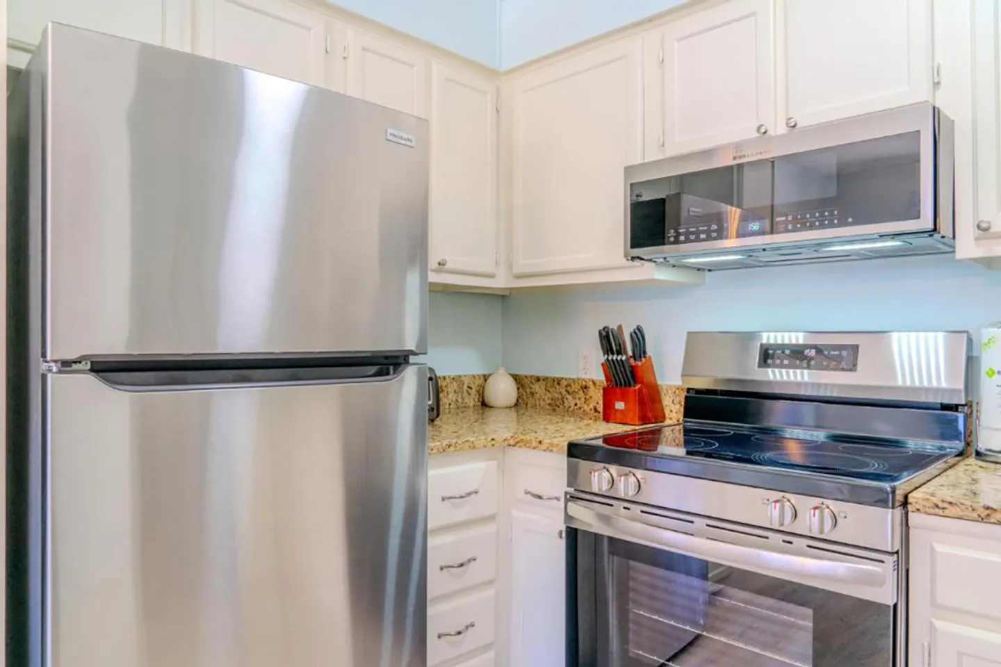 Kitchen corner with stainless steel refrigerator, stove, microwave, white cabinetry, and granite countertops.