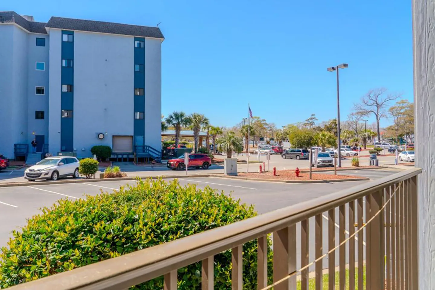 View from an outdoor balcony overlooking a parking lot, landscaped bushes, and adjacent multi-story residential buildings under a clear blue sky.