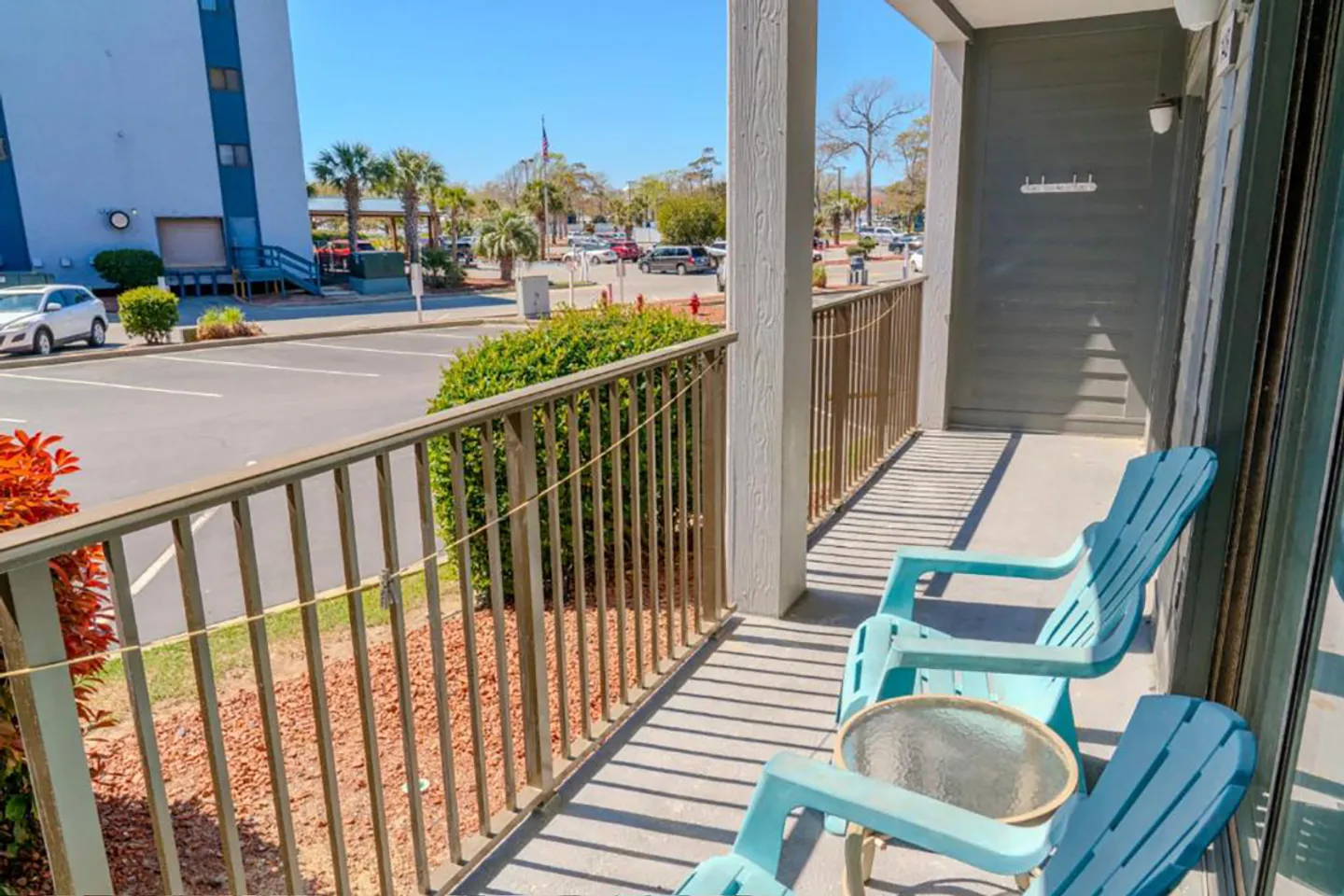 A covered balcony with two blue chairs and a small table overlooking a parking lot on a sunny day.