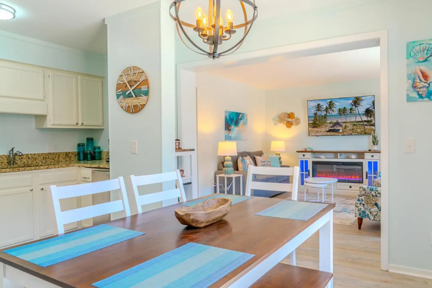 Dining area with a wooden tabletop, white chairs, and an orb chandelier, looking into the adjacent living room and kitchen.