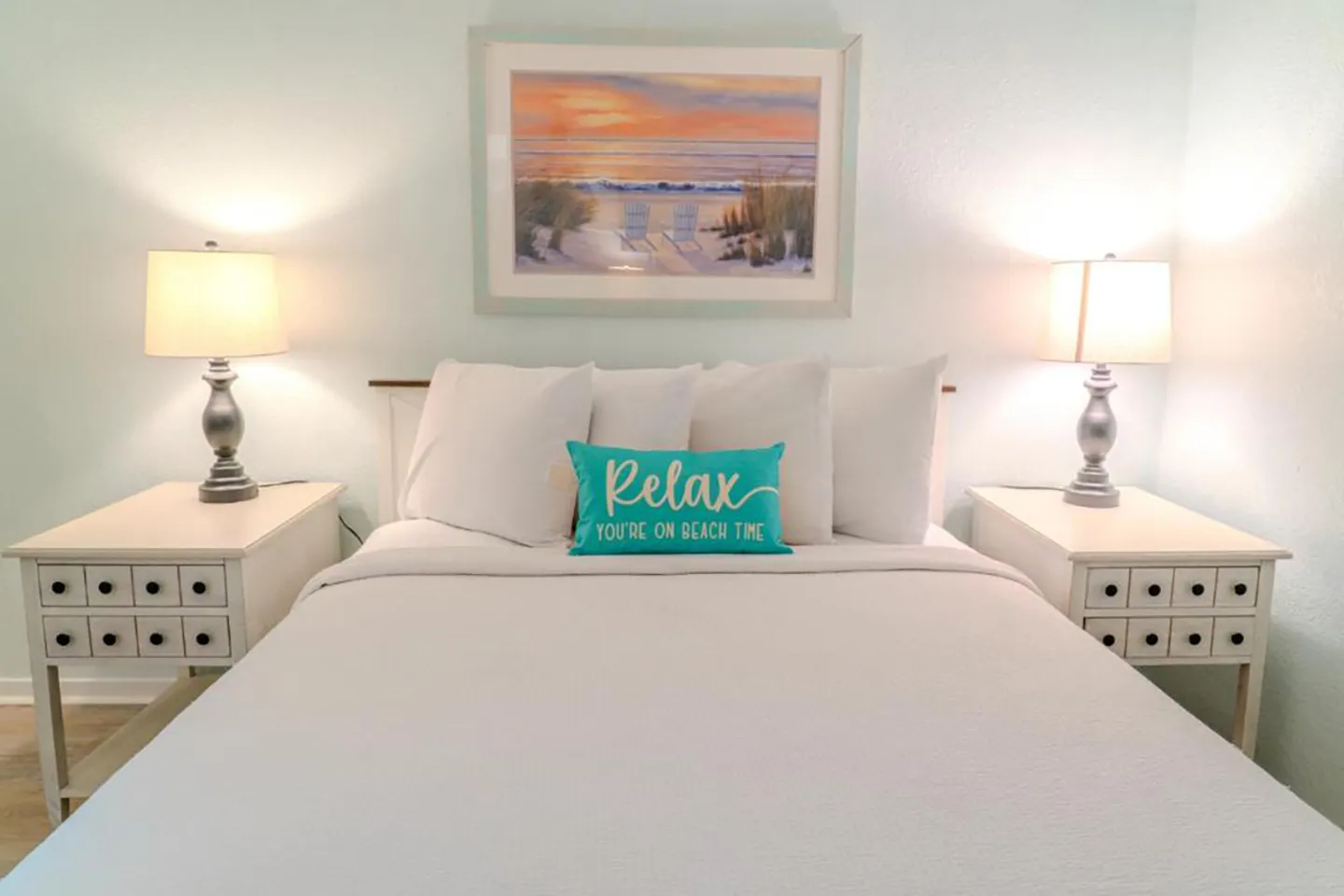 Bedroom with a made bed, white nightstands with lamps on each side, and beach-themed framed wall art.