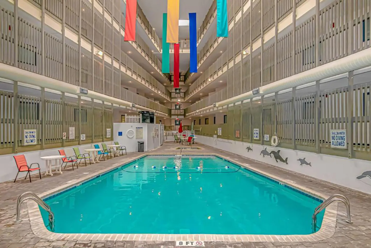 Indoor rectangular swimming pool located in a multi-story atrium surrounded by guest hallways and colorful banners hanging overhead.