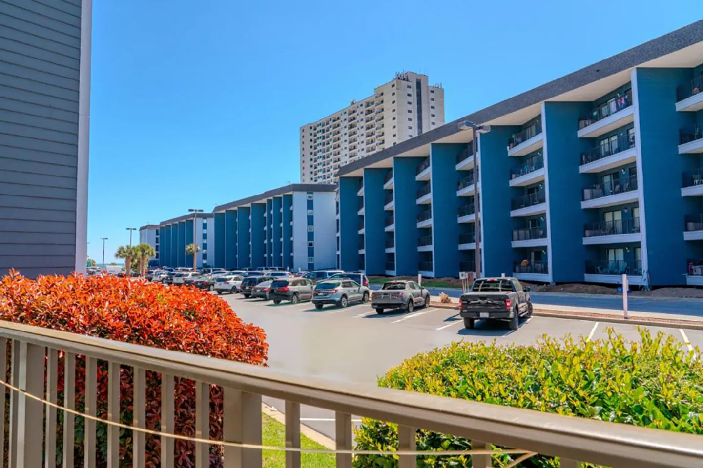 View from an outdoor balcony overlooking a parking lot and multi-story apartment buildings on a clear day.