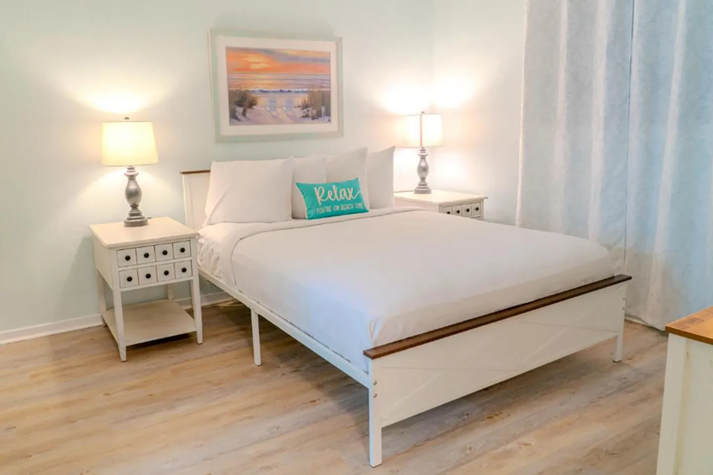 Bedroom with a white bed frame, matching white nightstands with lamps, wood-style flooring, and a beach artwork above the bed.