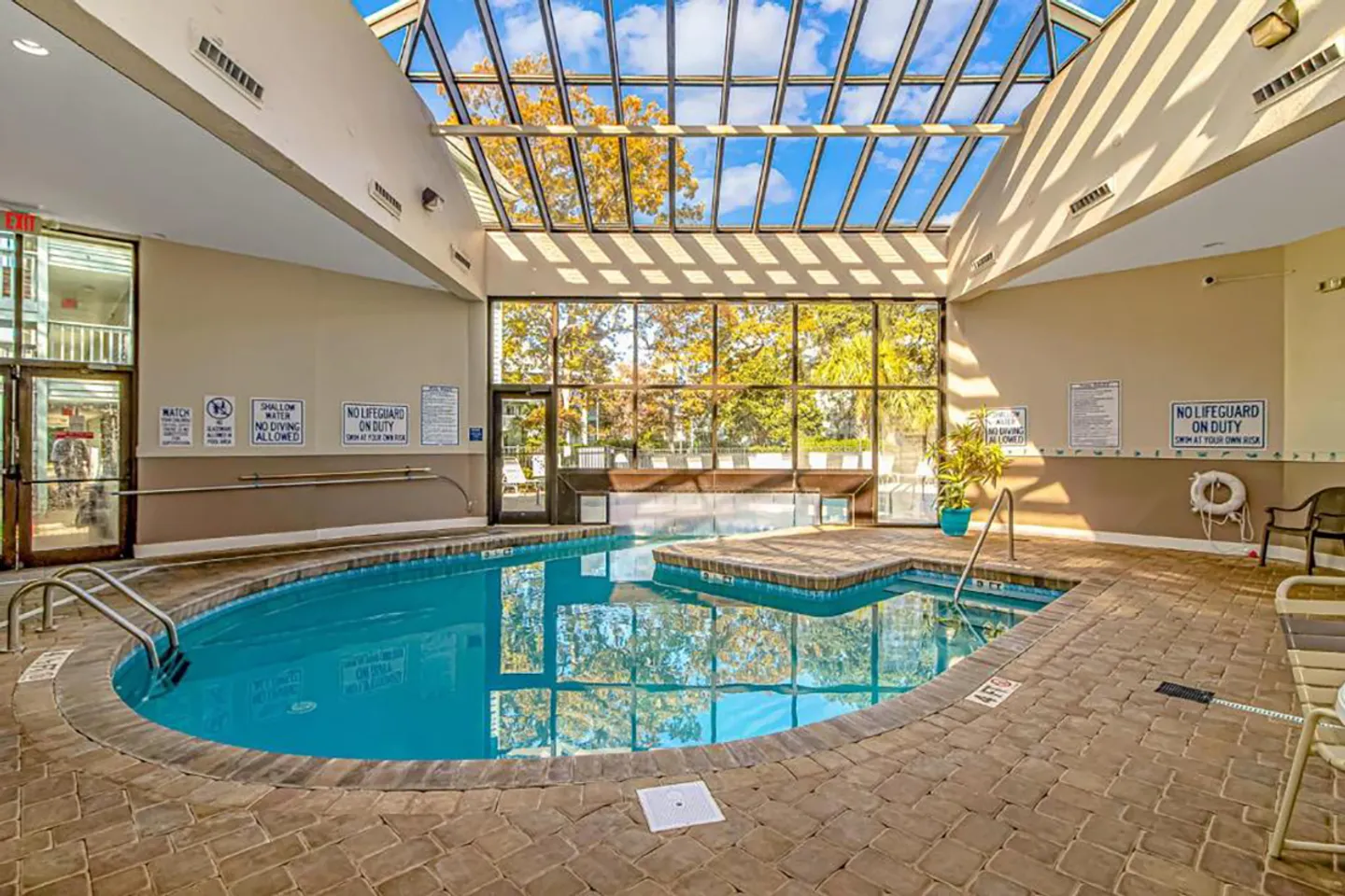 Indoor swimming pool surrounded by paved decking beneath a large glass skylight ceiling and floor-to-ceiling windows.