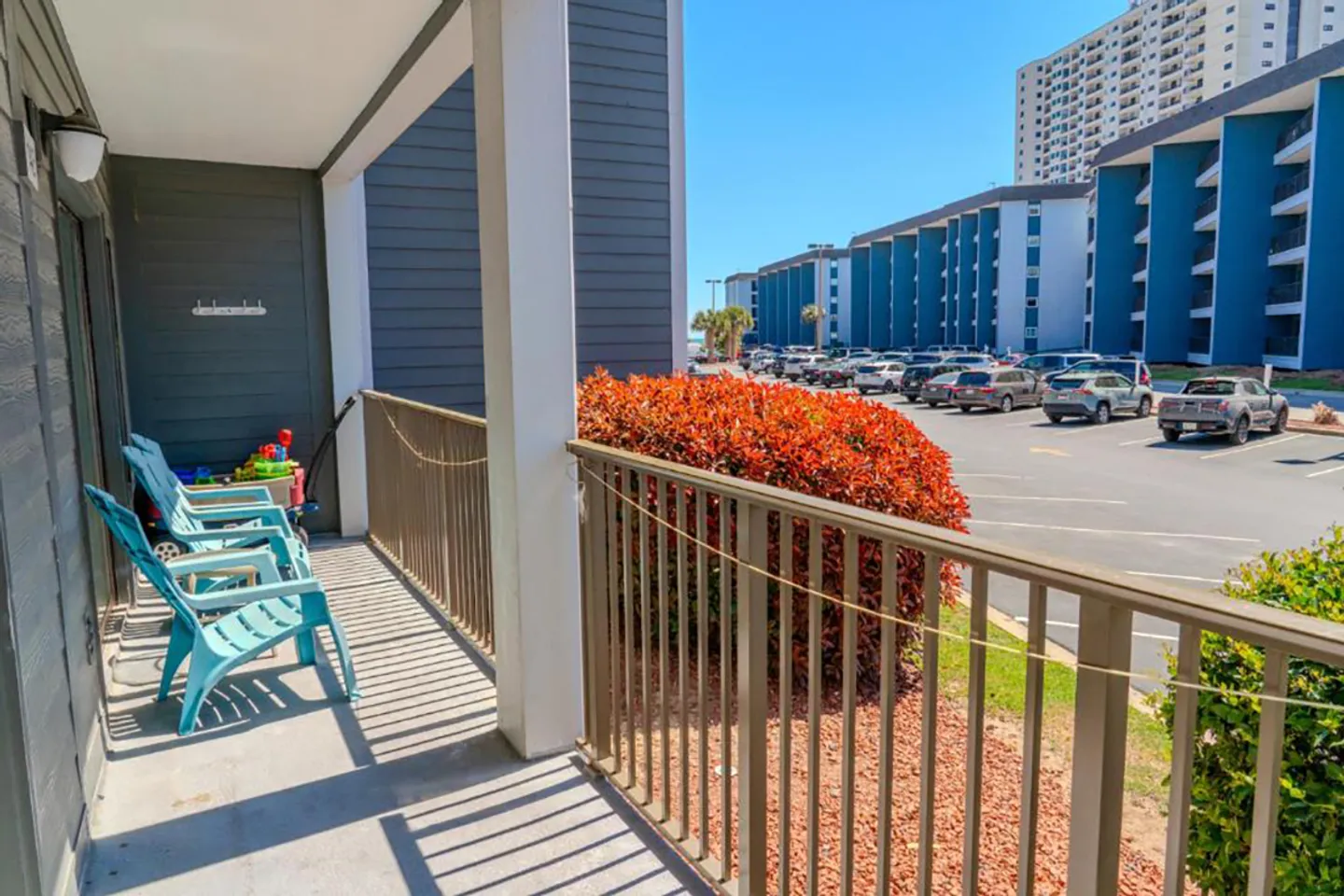 Covered patio with metal railings and turquoise outdoor chairs looking out toward a parking lot and condo buildings.