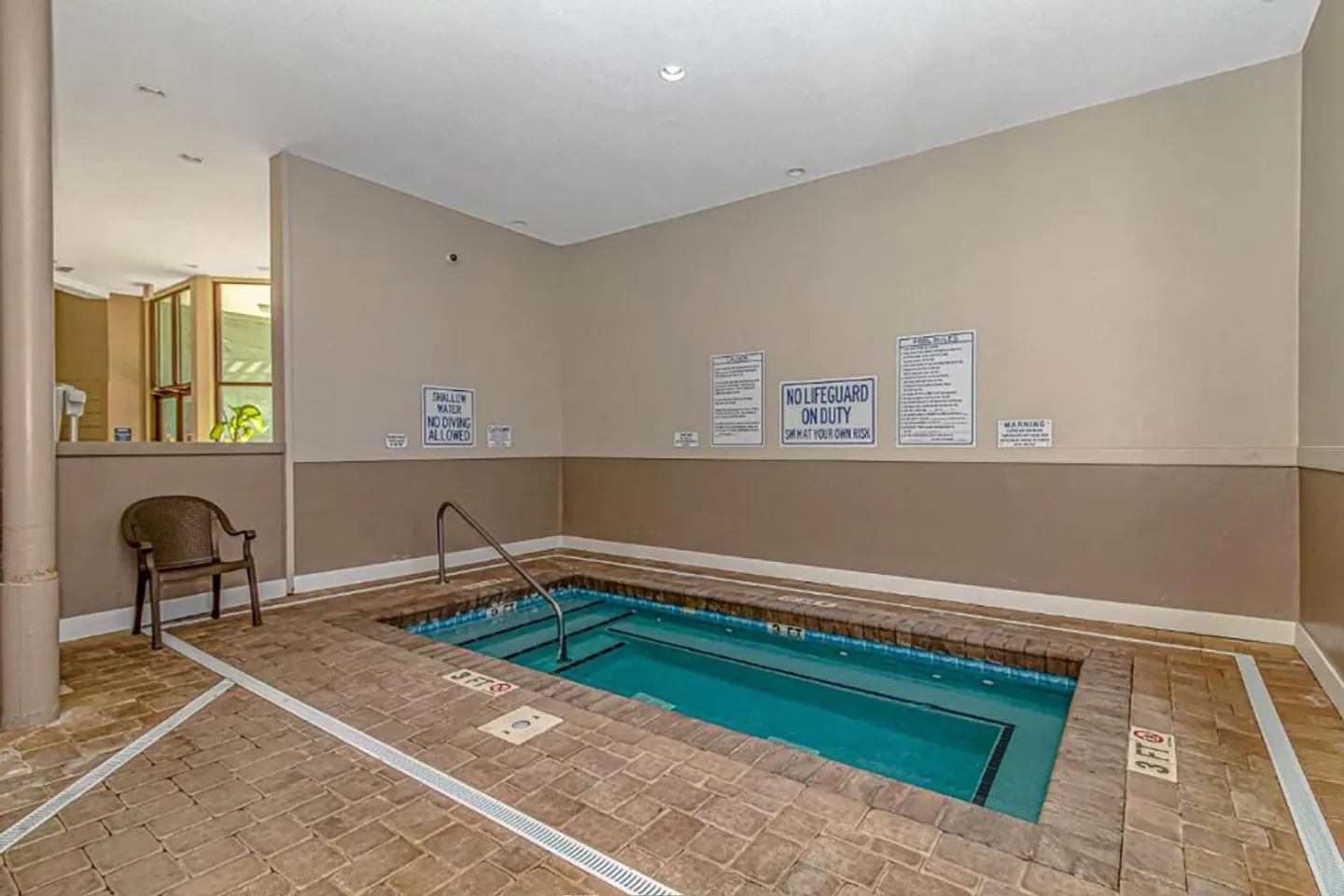 Indoor rectangular hot tub set into a paver deck with safety handrail and posted pool rules.
