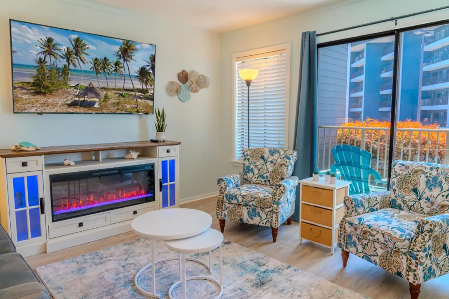 Living room featuring two floral armchairs, an electric fireplace credenza with a mounted TV, round nesting coffee tables, and a sliding glass door to a balcony.