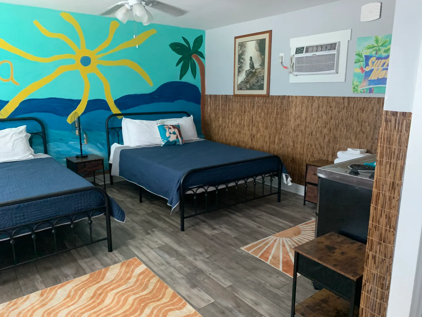 Bedroom featuring two black metal frame beds, a vibrant beach-themed mural, wood-look flooring, and a wall-mounted air conditioner.