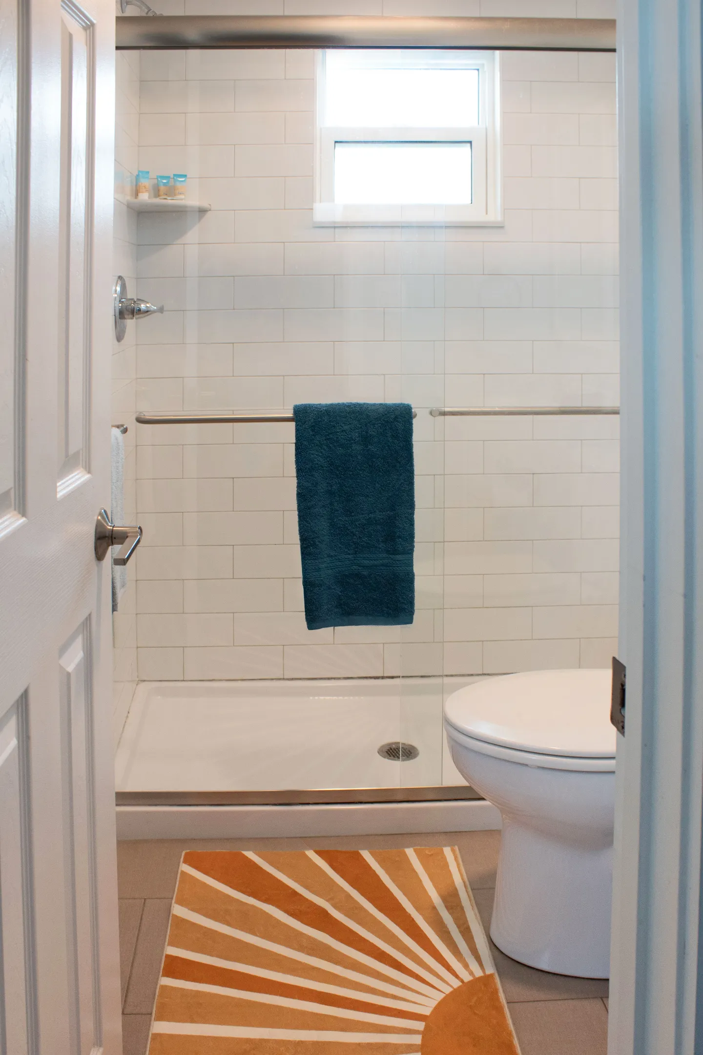 Bathroom view through an open door showing a toilet, a shower stall with sliding glass doors, white subway tile, and a patterned floor mat.