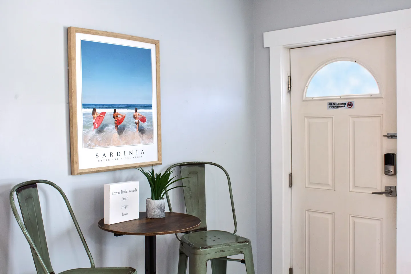 Entryway corner featuring a framed beach poster above a small table and green metal chairs next to a white front door.