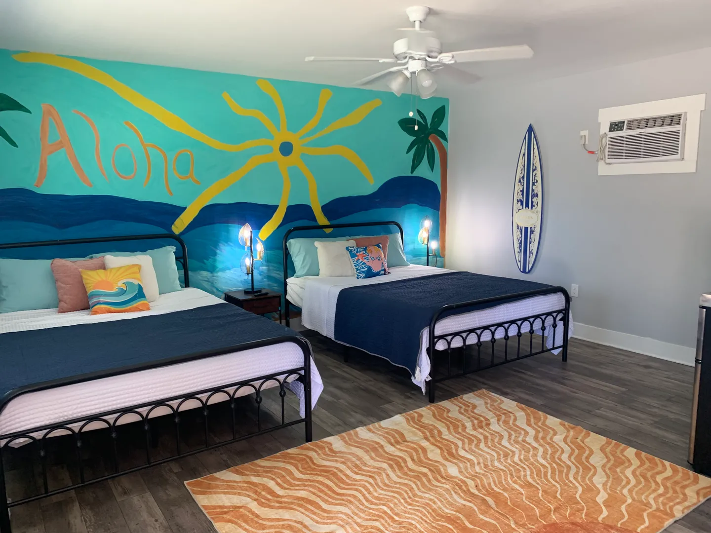 Bedroom featuring two beds with black metal frames, an orange patterned rug, and a hand-painted beach-themed wall mural that reads Aloha.