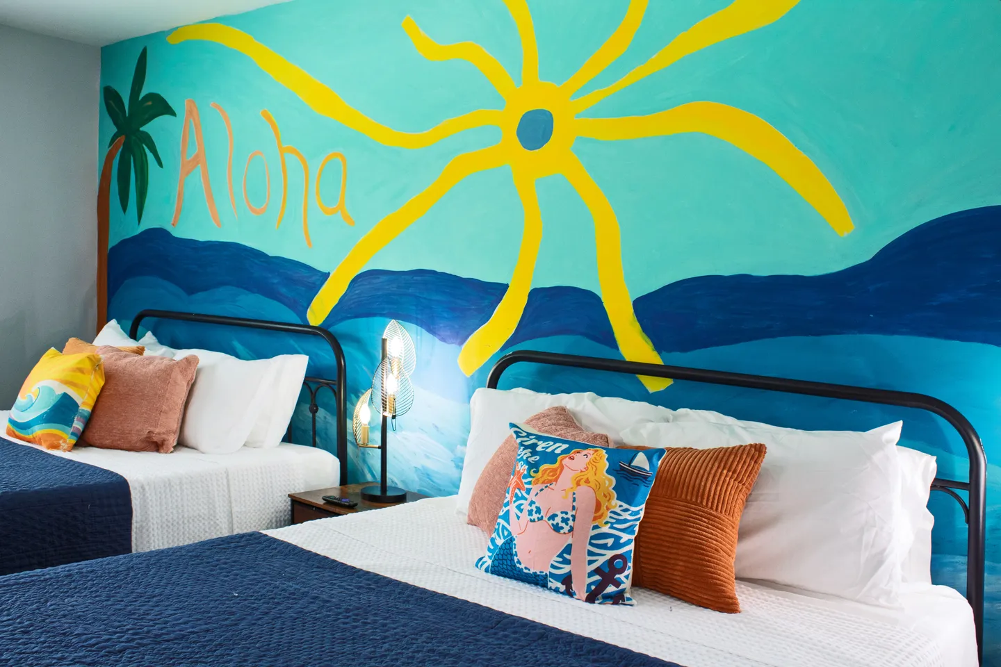Bedroom featuring two beds with black metal frames set against a colorful wall mural depicting a beach scene with a bright yellow sun, turquoise water, and the word Aloha.
