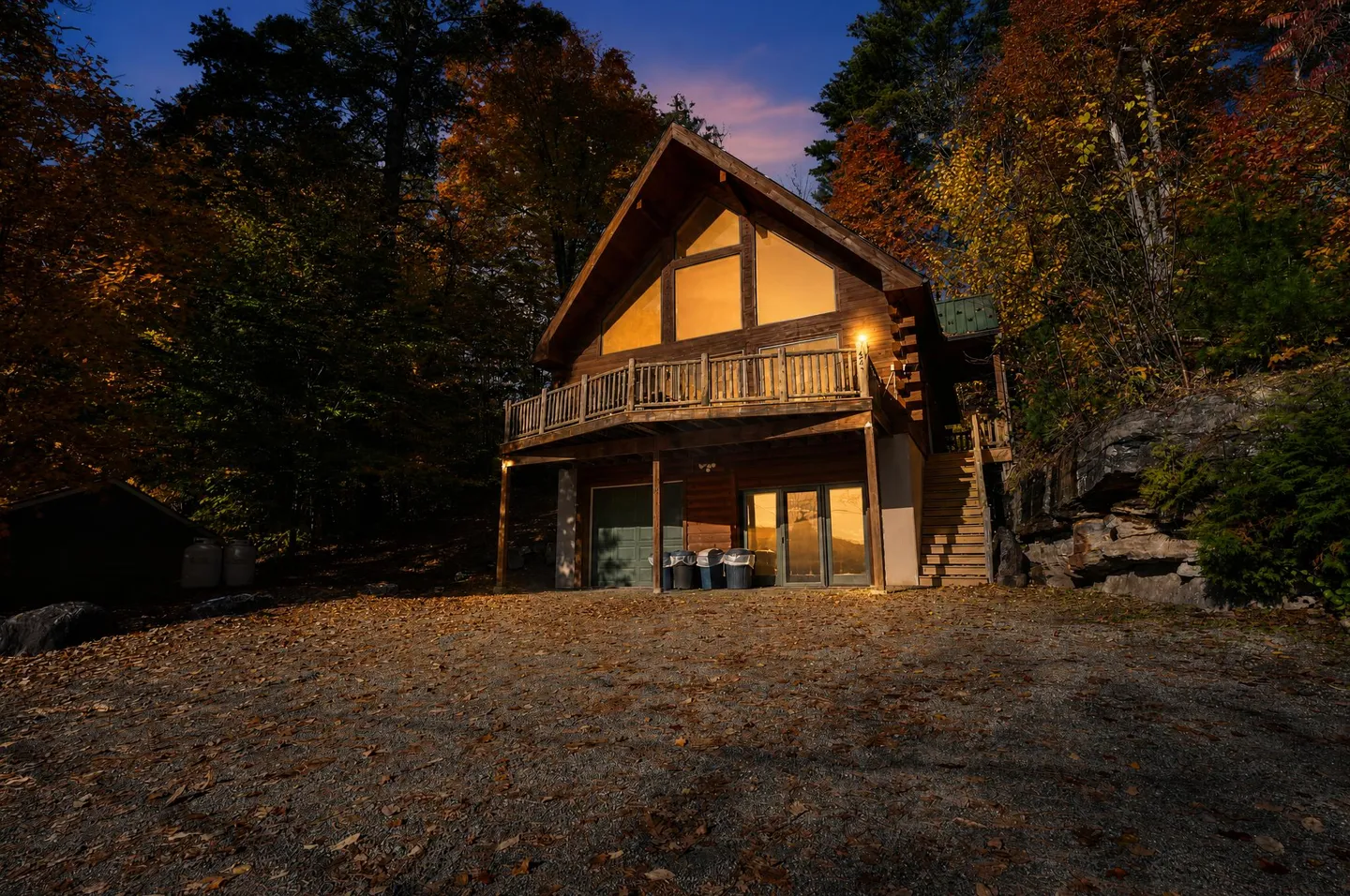 Exterior dusk view of an A-frame wooden cabin nestled in the forest with glowing windows, an upper deck, and gravel parking area.