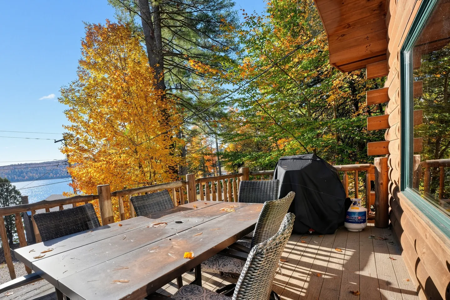 Outdoor wooden deck of a log home featuring a dining table with wicker chairs, a covered barbecue grill, and autumn trees overlooking a body of water.