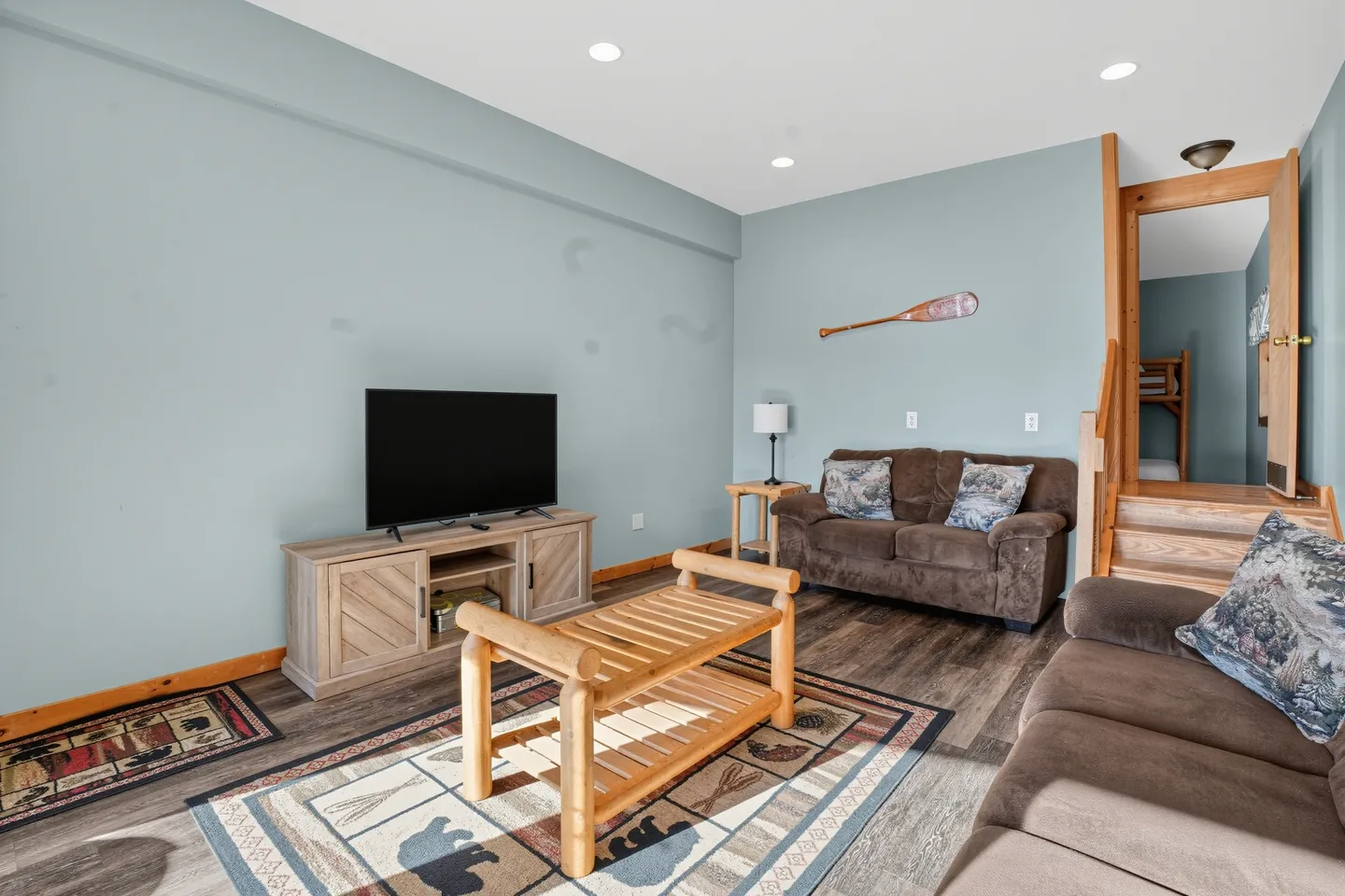 Living room with light blue walls, brown sofas, a wooden coffee table, a TV on a console, and stairs leading to another room.