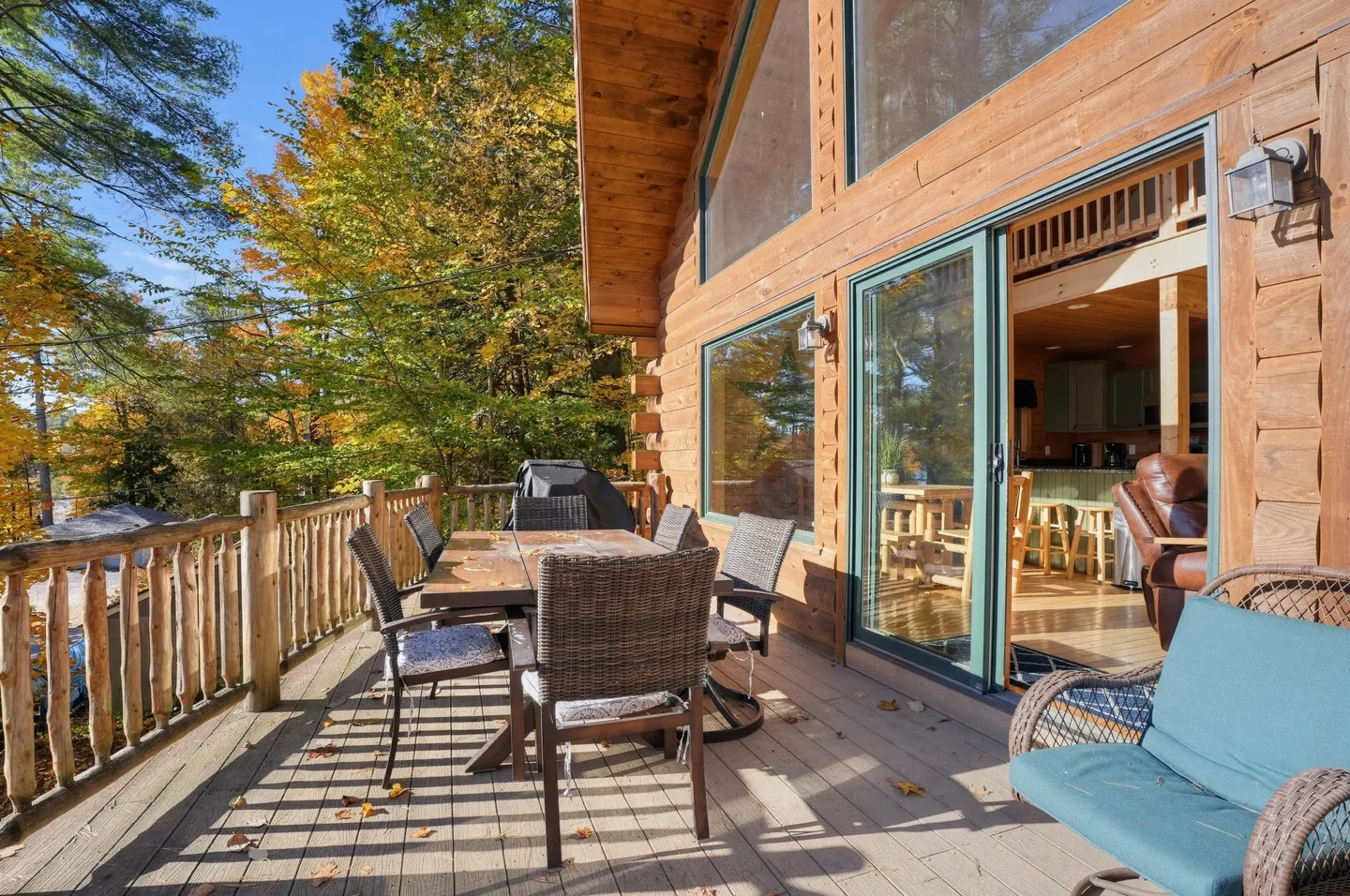 A wooden deck with an outdoor dining table and chairs along the exterior of a log home surrounded by autumn trees.