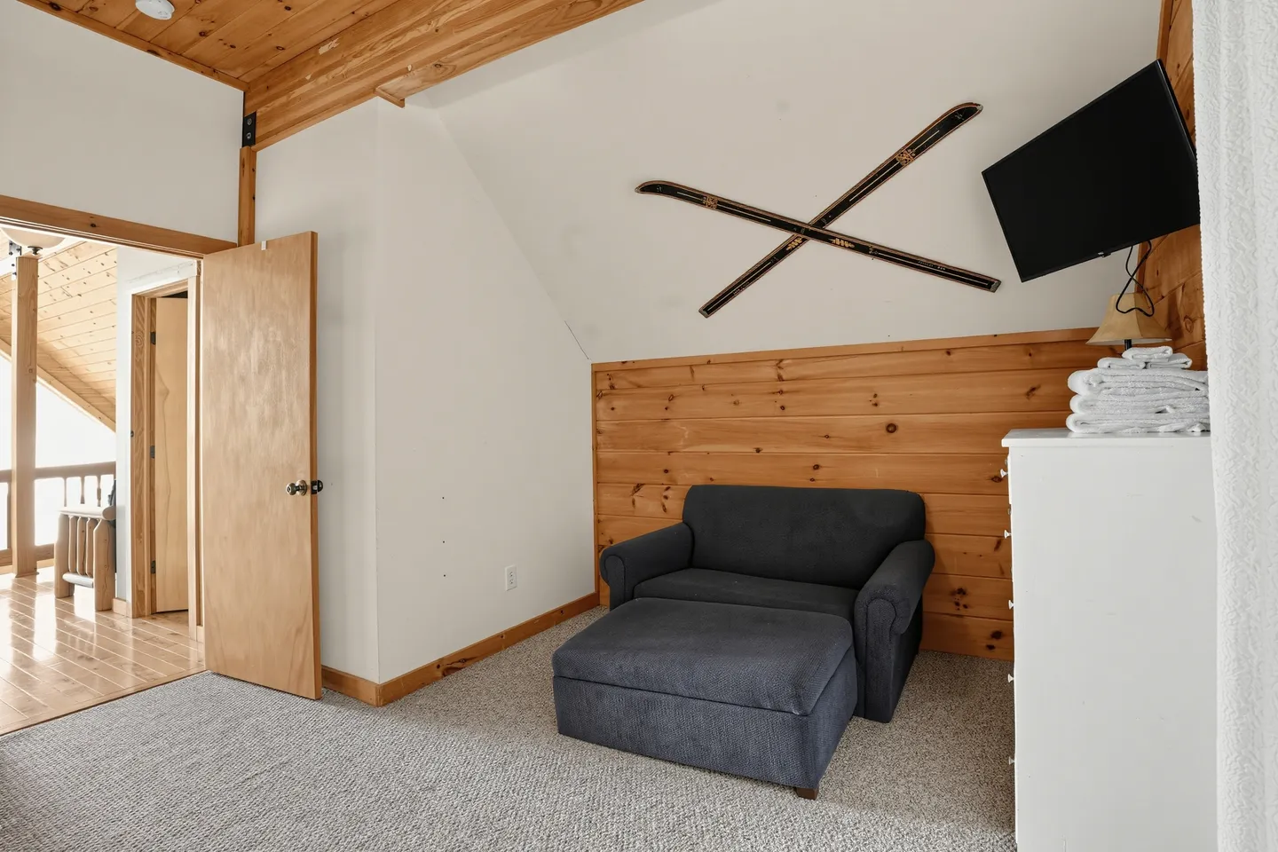 Carpeted room featuring an upholstered armchair with an ottoman, wood-paneled accents, decorative crossed skis on the wall, and an open doorway.