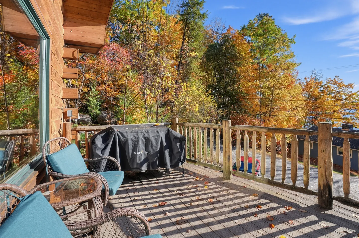 Outdoor wooden deck of a log home with log railings, patio chairs, a covered grill, and vibrant autumn trees in the background.