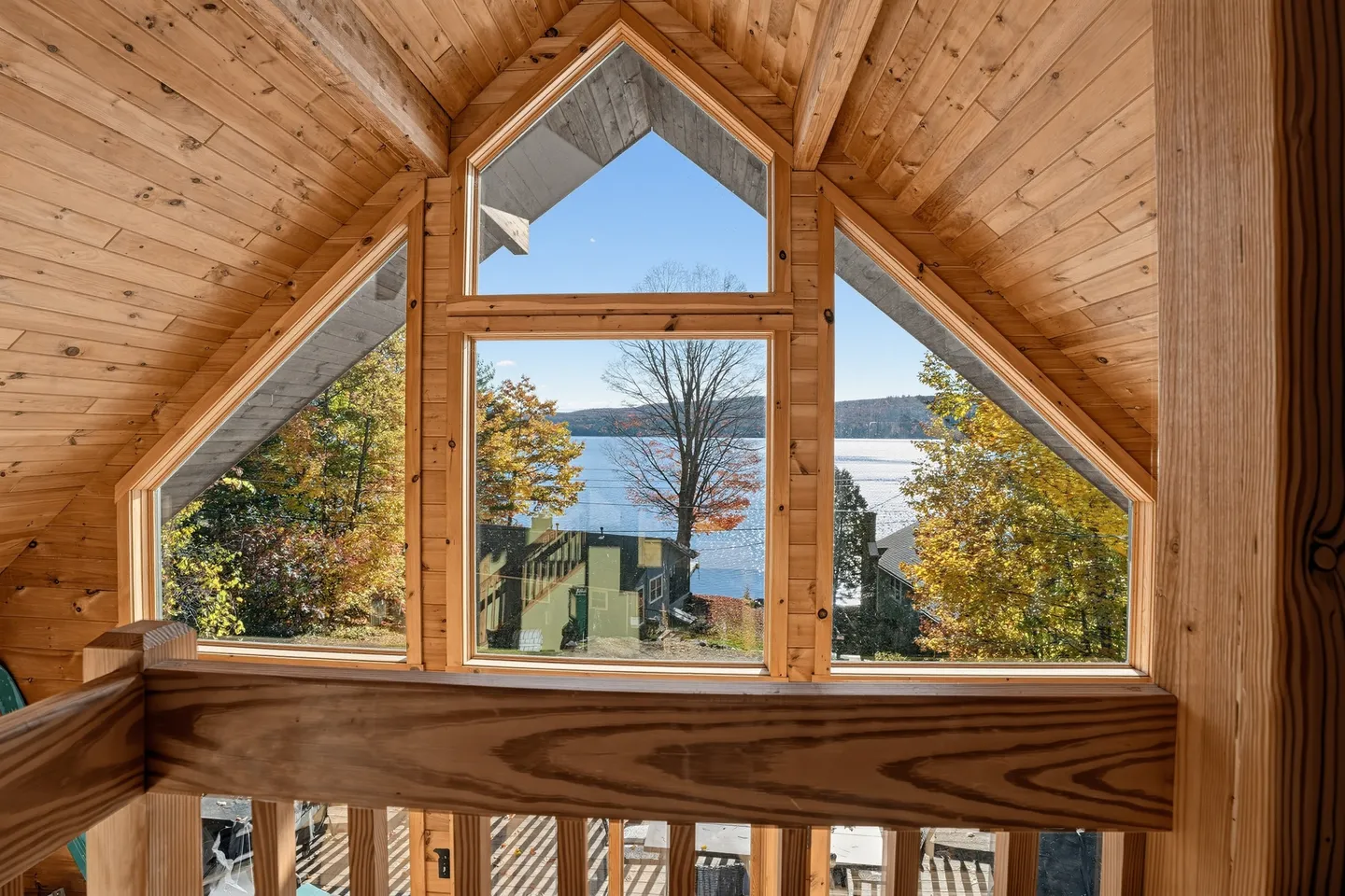 Large picture windows framed by natural wood under a vaulted wood-plank ceiling overlook a lake and autumn trees.