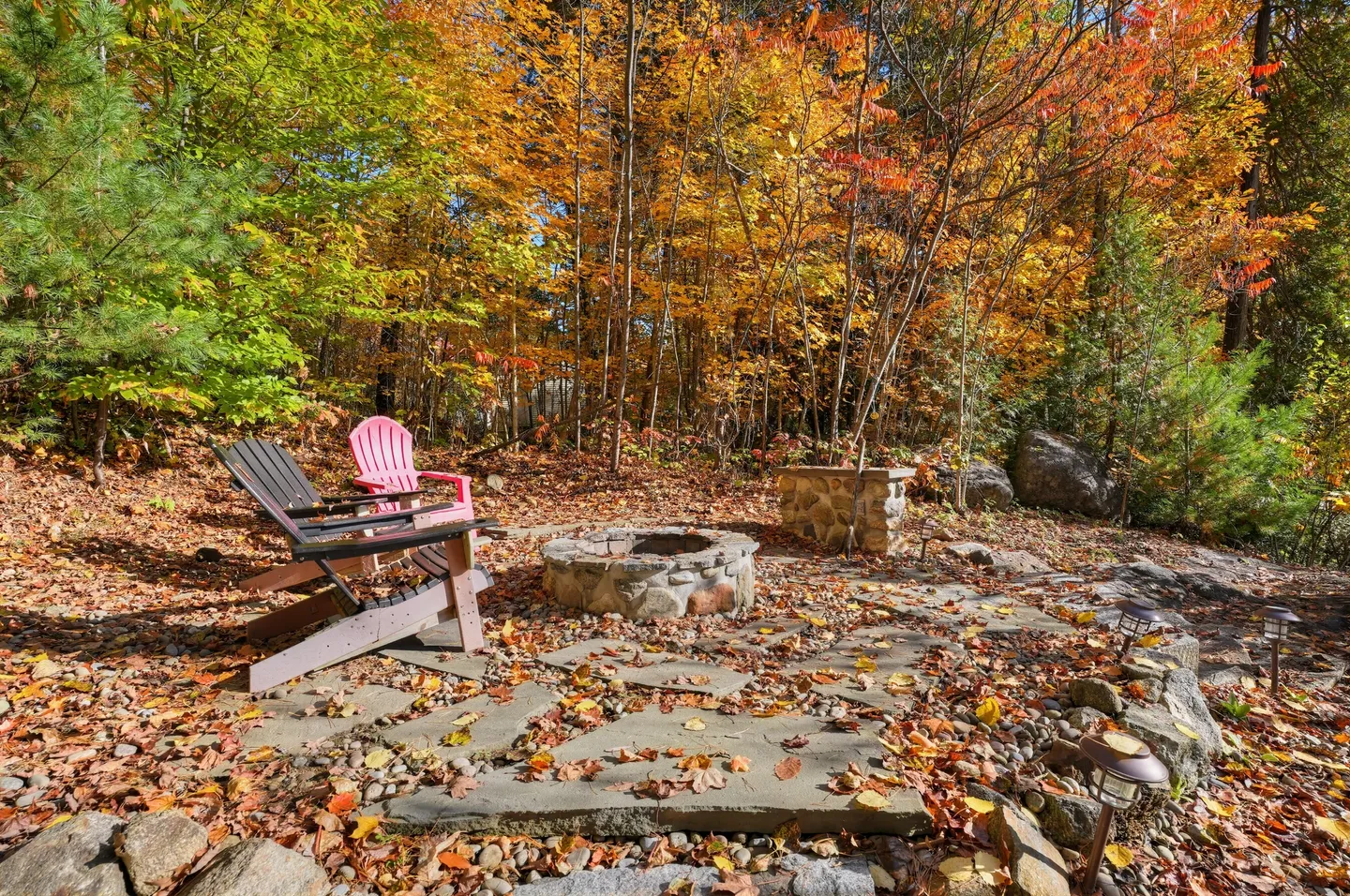 Outdoor stone fire pit with Adirondack chairs on a flagstone patio surrounded by fallen autumn leaves and dense woods.