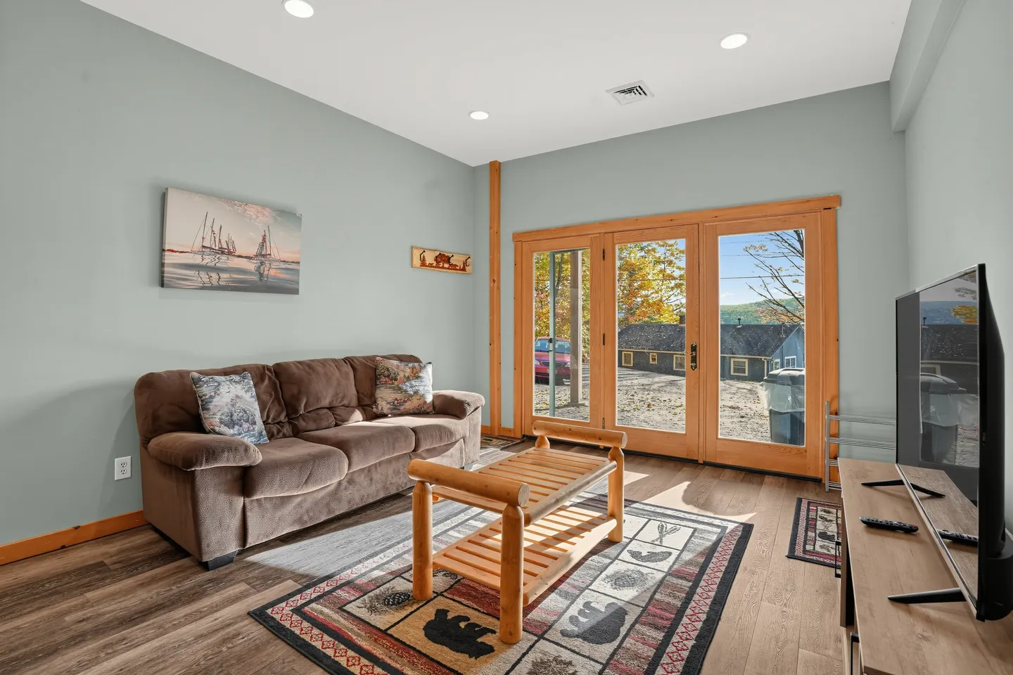 Living room with a brown sofa, log-style coffee table, area rug with nature motifs, and large glass-paned doors leading outside.