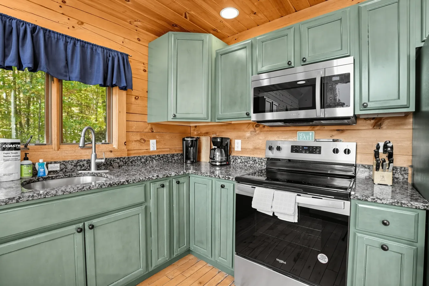 Kitchen in a log cabin with sage green cabinets, dark speckled countertops, stainless steel appliances, and a window.