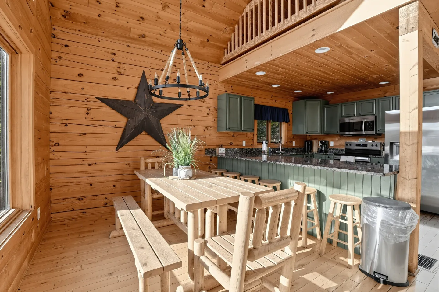 Dining area with a wooden log-style table, benches, and chairs adjacent to a kitchen with green cabinets and a breakfast bar.