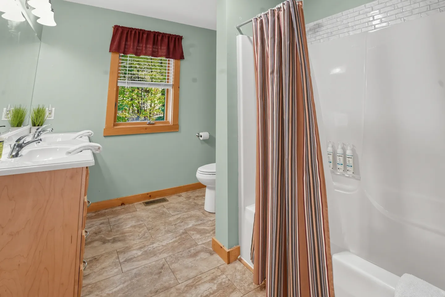 Bathroom with a double vanity sink, toilet, window with red valance, and combination bathtub shower with a striped curtain.