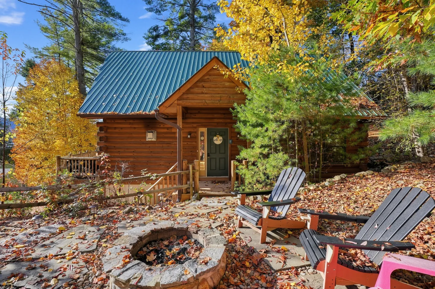 Exterior view of a log cabin with a green metal roof, stone fire pit, and Adirondack chairs surrounded by autumn foliage.