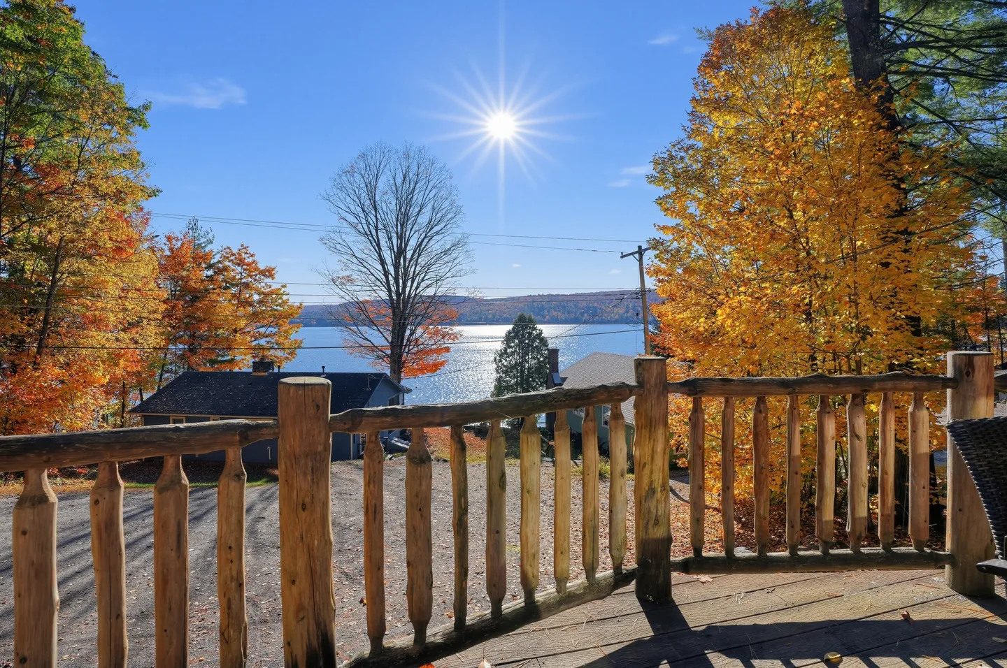 Elevated wooden deck with log railings overlooking trees in autumn foliage, neighboring roofs, and a sunlit lake under a clear blue sky.