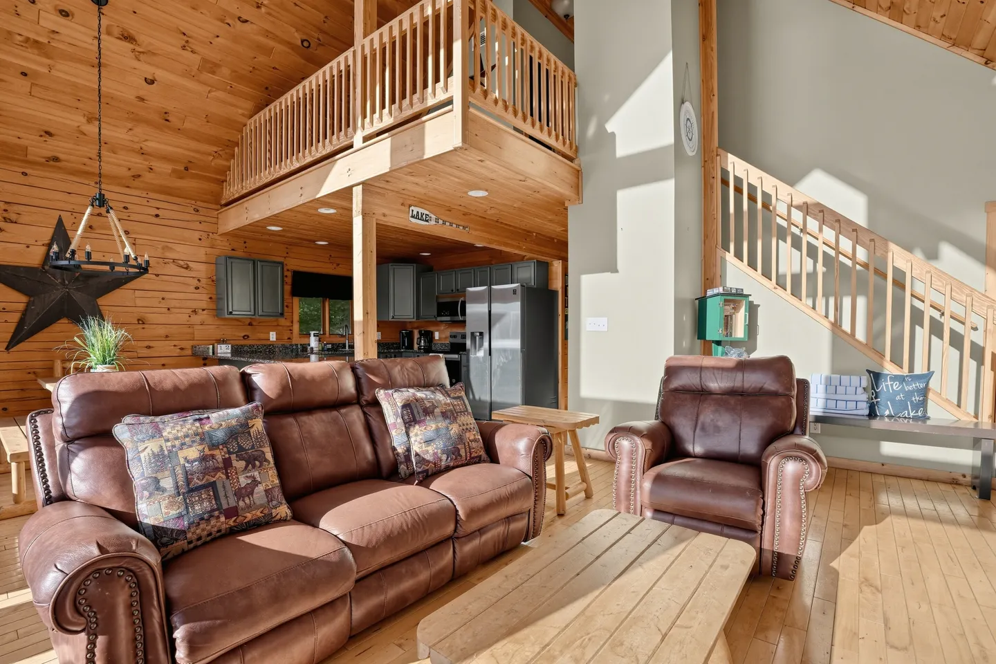 Living room with brown leather seating, wood-paneled walls, an open loft above, and a kitchen visible in the background.
