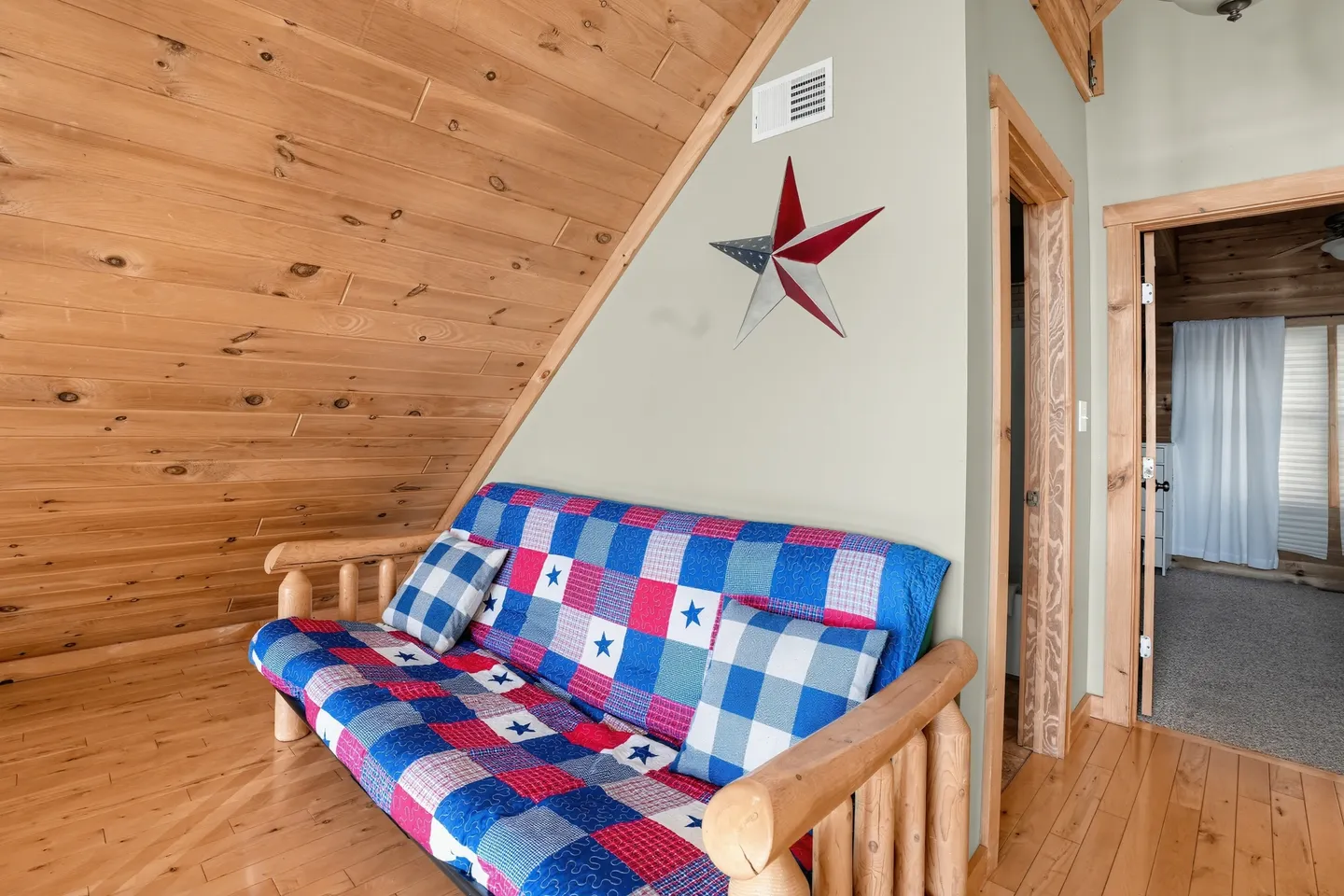 A futon with a red, white, and blue patterned quilt beneath a slanted wood-paneled ceiling.