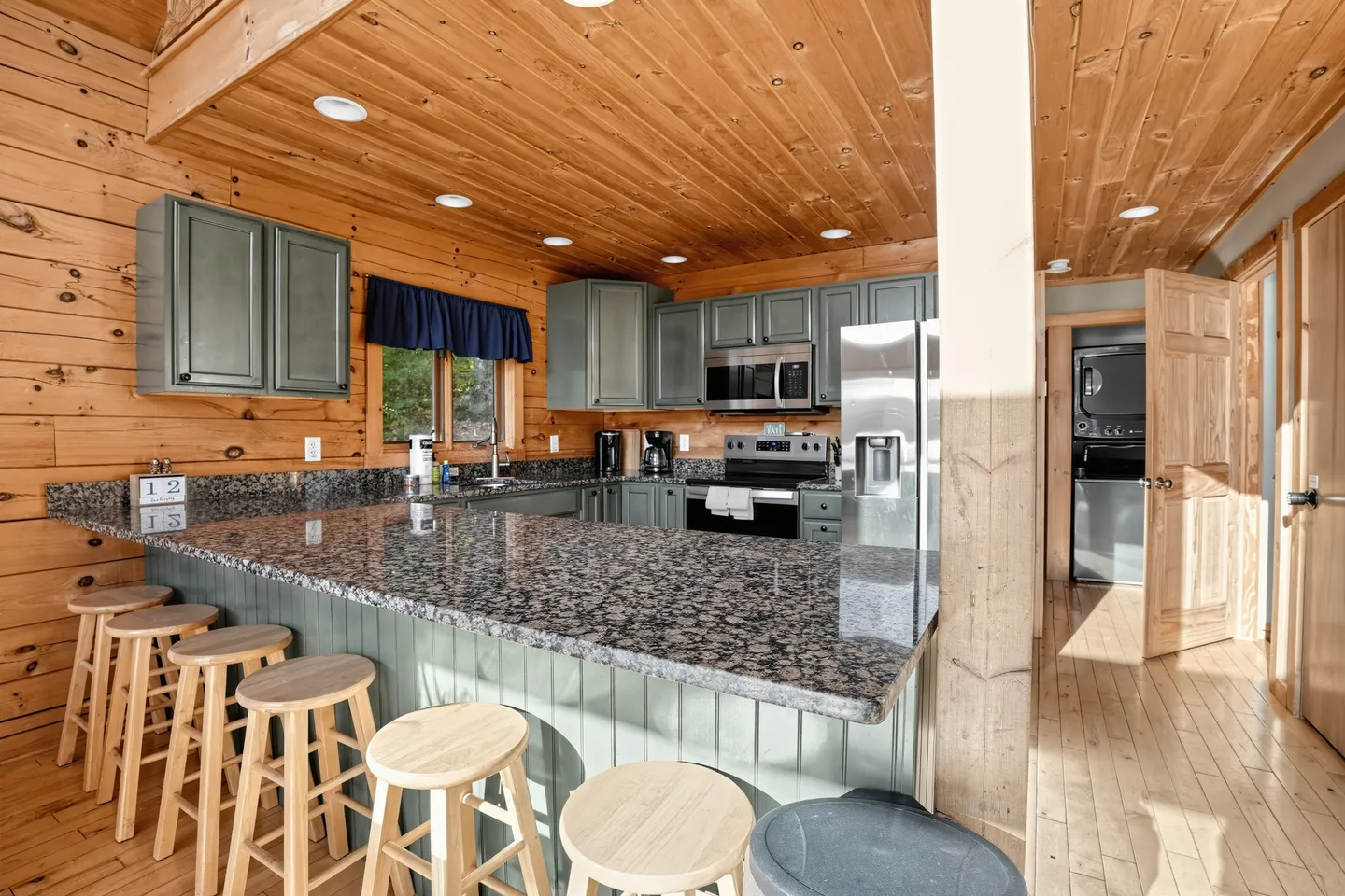 Kitchen in a log-style home with a curved granite-topped peninsula, wooden barstools, painted green cabinetry, and stainless steel appliances.