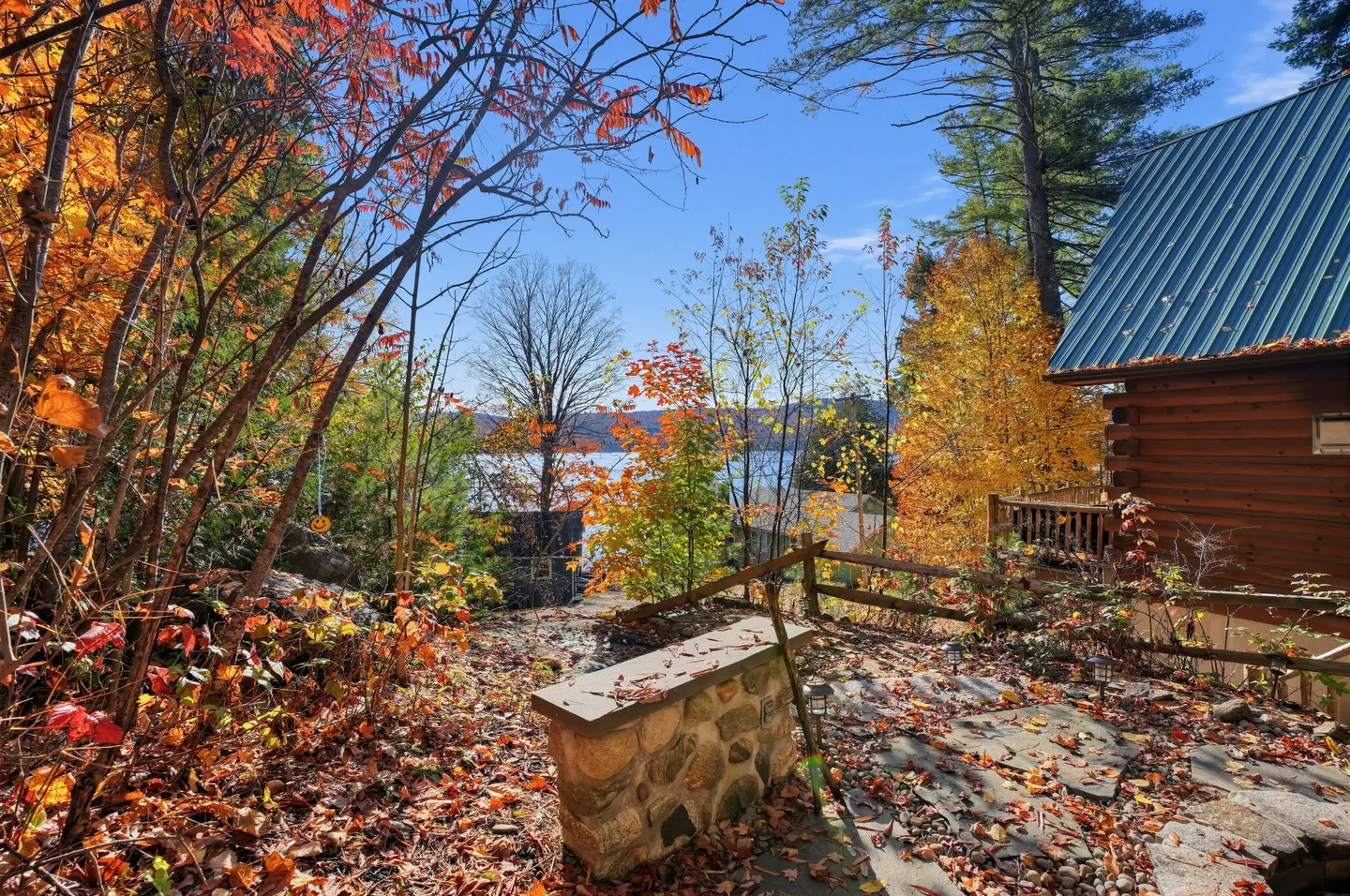 Outdoor stone patio covered in autumn leaves next to a log cabin, overlooking trees and a lake under a clear blue sky.