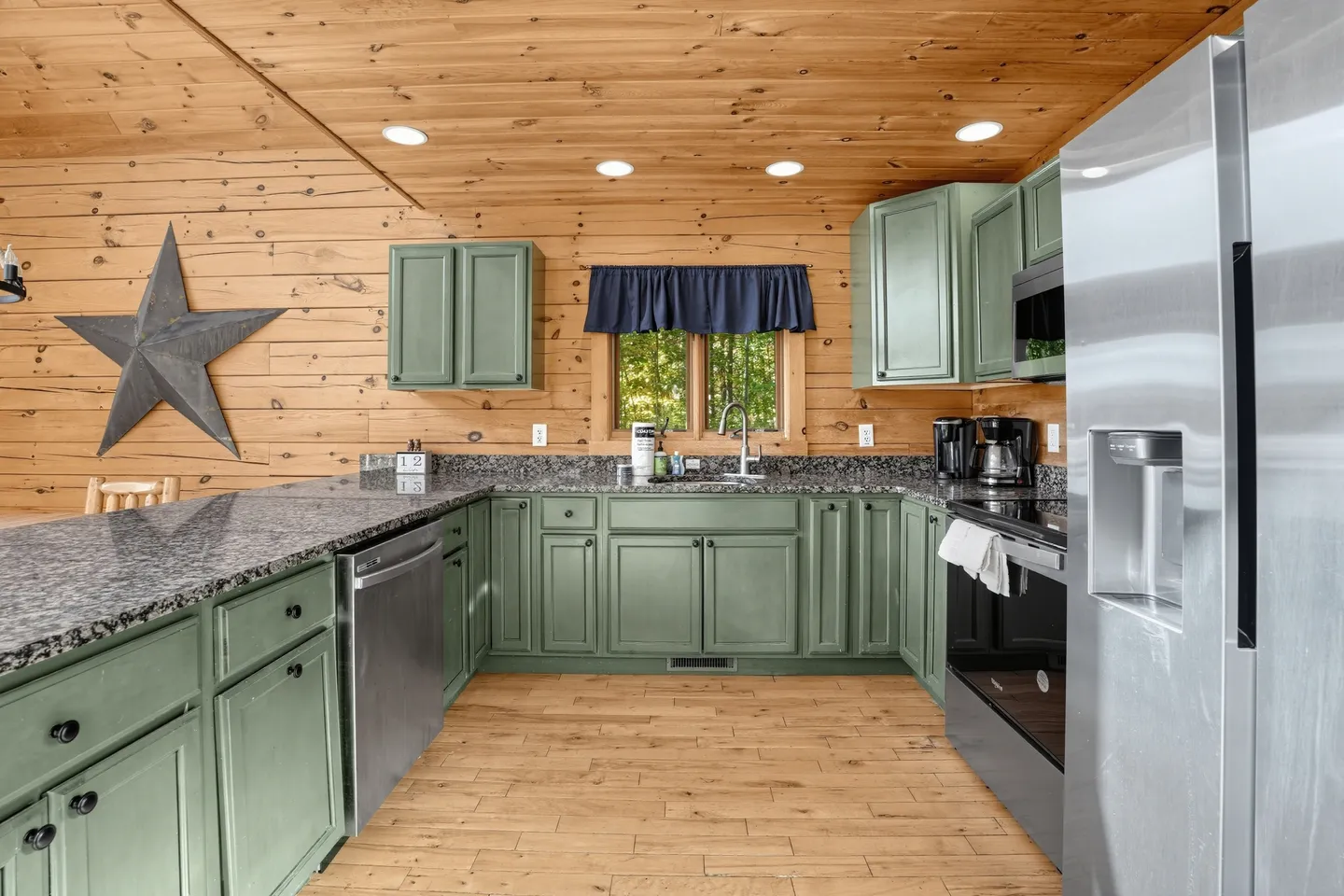 Kitchen with wood-paneled walls and ceiling, sage green cabinetry, dark granite-look countertops, and stainless steel appliances.