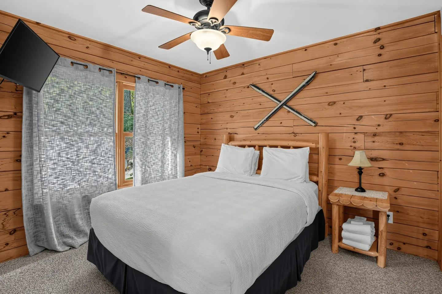 Bedroom with wood-paneled walls, a log bed frame with white bedding, a ceiling fan, and decorative skis on the wall.