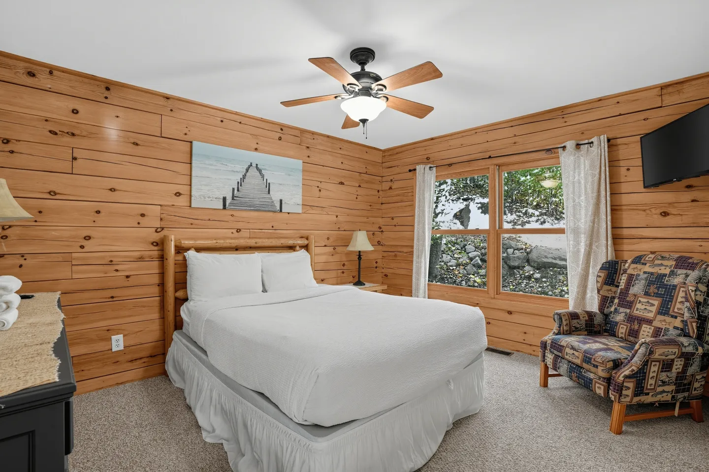Bedroom with wood-paneled walls, a bed made with white linens, a ceiling fan, and an armchair next to a window.