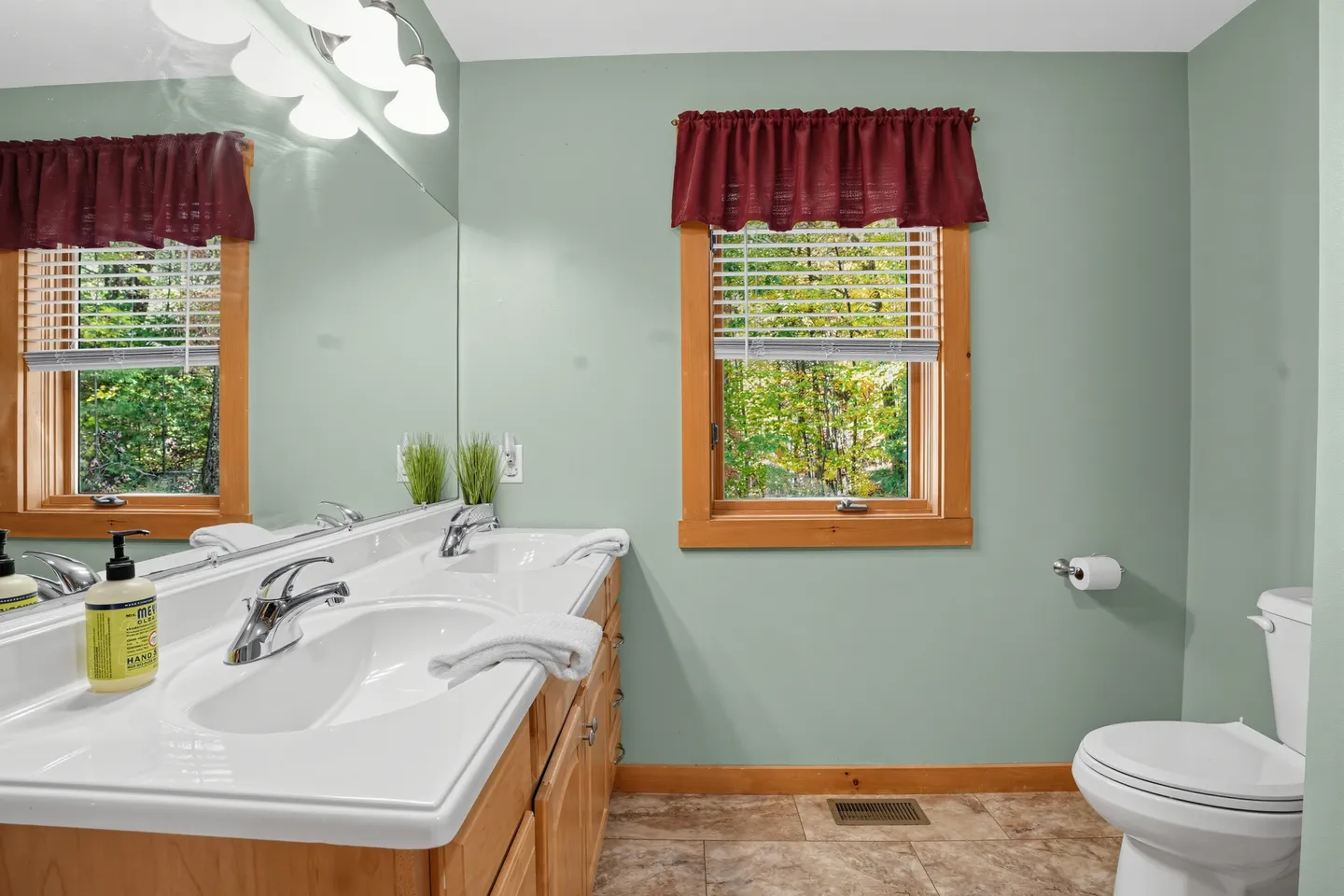 Bathroom with a double-sink wood vanity, large mirror, green walls, a toilet, and a window with wood trim.