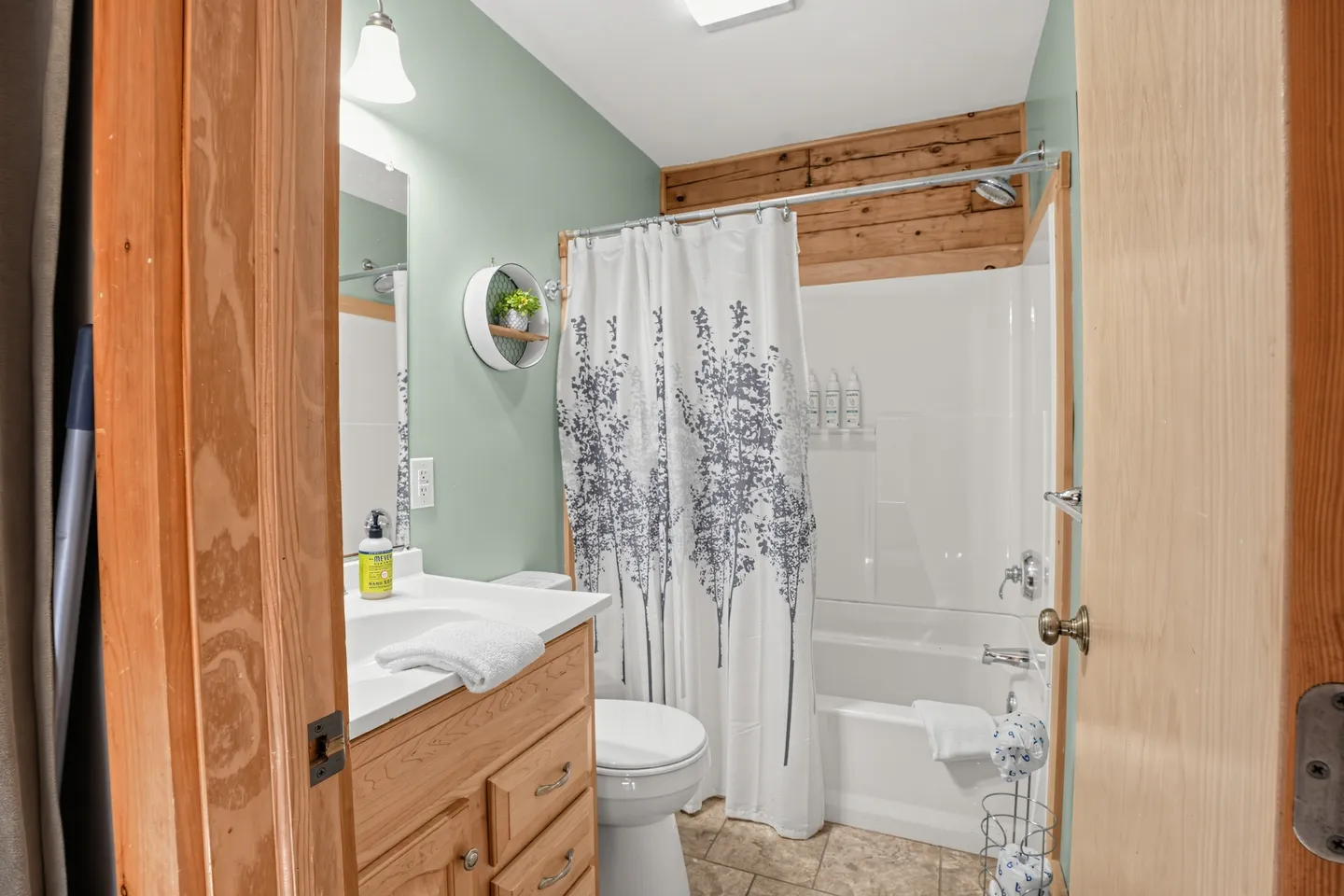 Bathroom with wooden vanity, toilet, and shower-tub combination featuring a tree-patterned curtain.