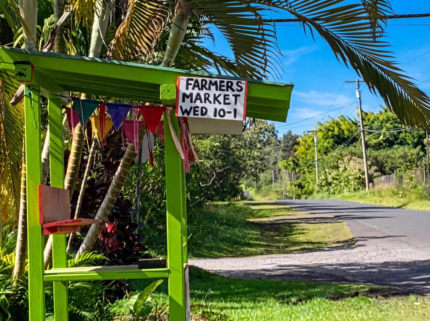 A lime-green roadside wooden stand decorated with colorful pennant banners and a sign reading Farmers Market Wed 10-1.