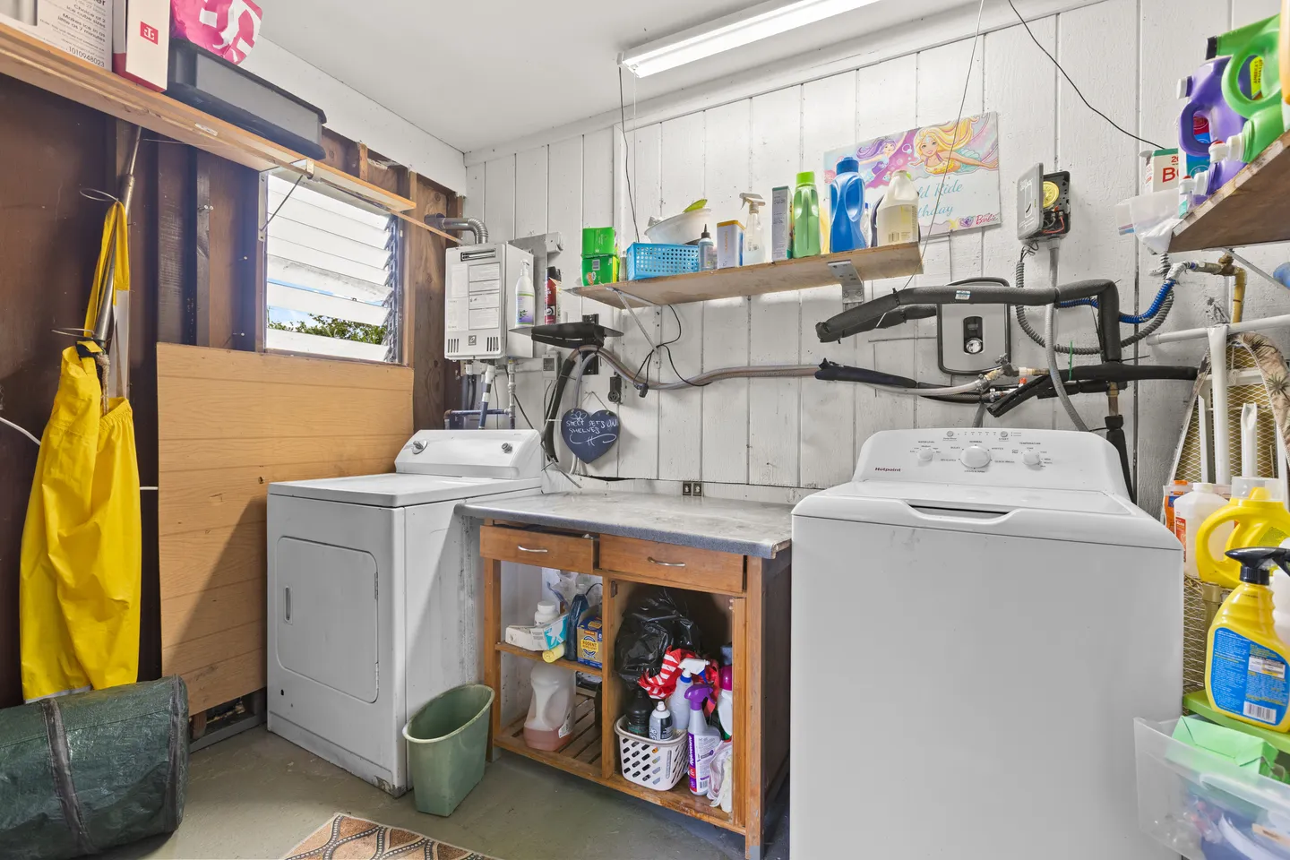 A laundry and utility area with a white washing machine, dryer, central wooden work table, and shelves holding cleaning supplies.