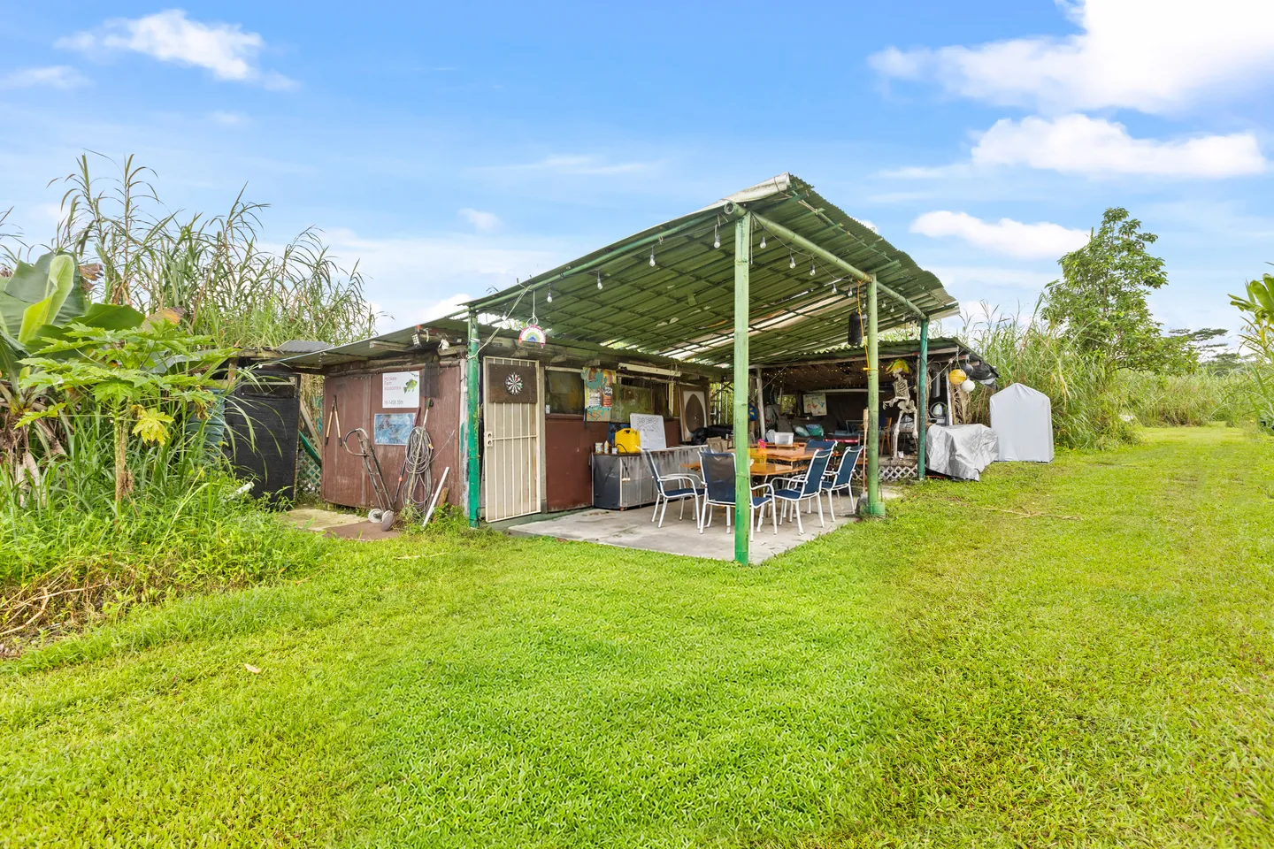 Exterior view of an outdoor covered patio structure with dining table and chairs surrounded by a grassy lawn and tropical foliage under a blue sky.