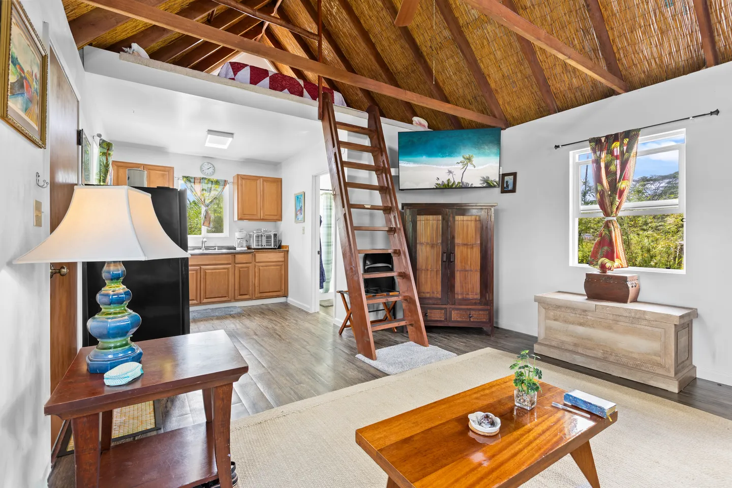 Open-concept living room and kitchen area featuring a thatched ceiling, wooden ladder to a loft, wall-mounted television, and coffee table.